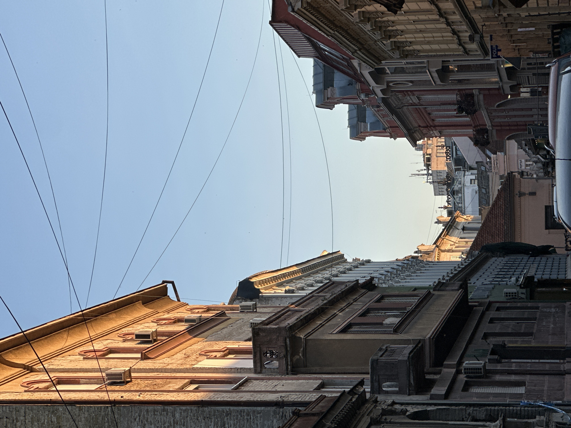 The image captures a street view in Novi Sad, Serbia, with buildings lining the street. The camera angle is tilted upwards, focusing on the architectural details of the buildings and the sky. Wires stretch across the sky, intersecting the buildings. The buildings exhibit various architectural styles, with different colors, materials, and details like windows, roofs, and air conditioning units. A vehicle is partially visible on the upper right side of the image. The sky is light blue.