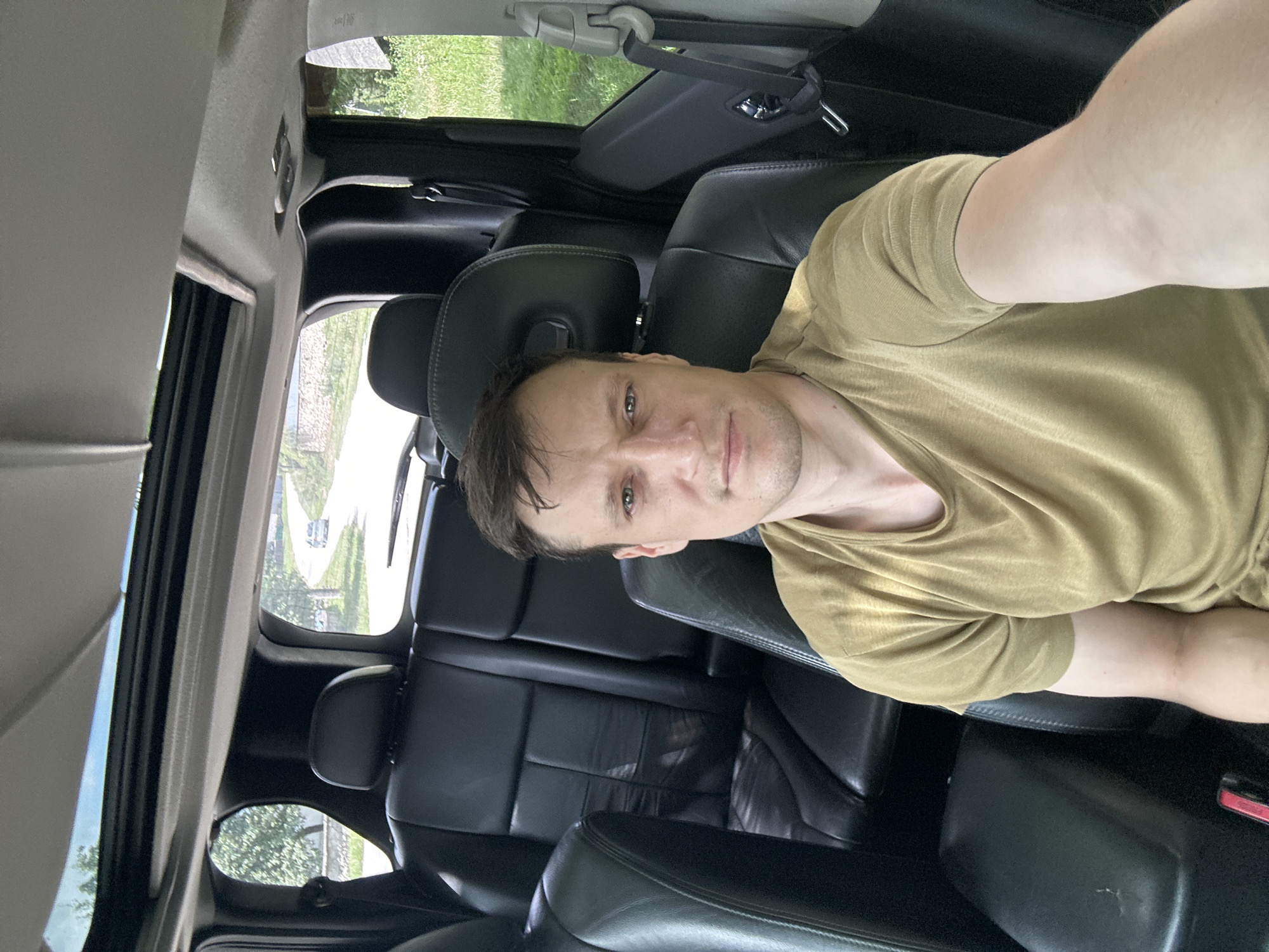 A man is taking a selfie inside a car. He is seated in the front passenger seat, looking directly at the camera. The car's interior is visible, with black leather seats and a sunroof open above. The background shows a rural road with some trees and greenery. The man is wearing a plain, olive-colored t-shirt. The location is Makiv, Ukraine.