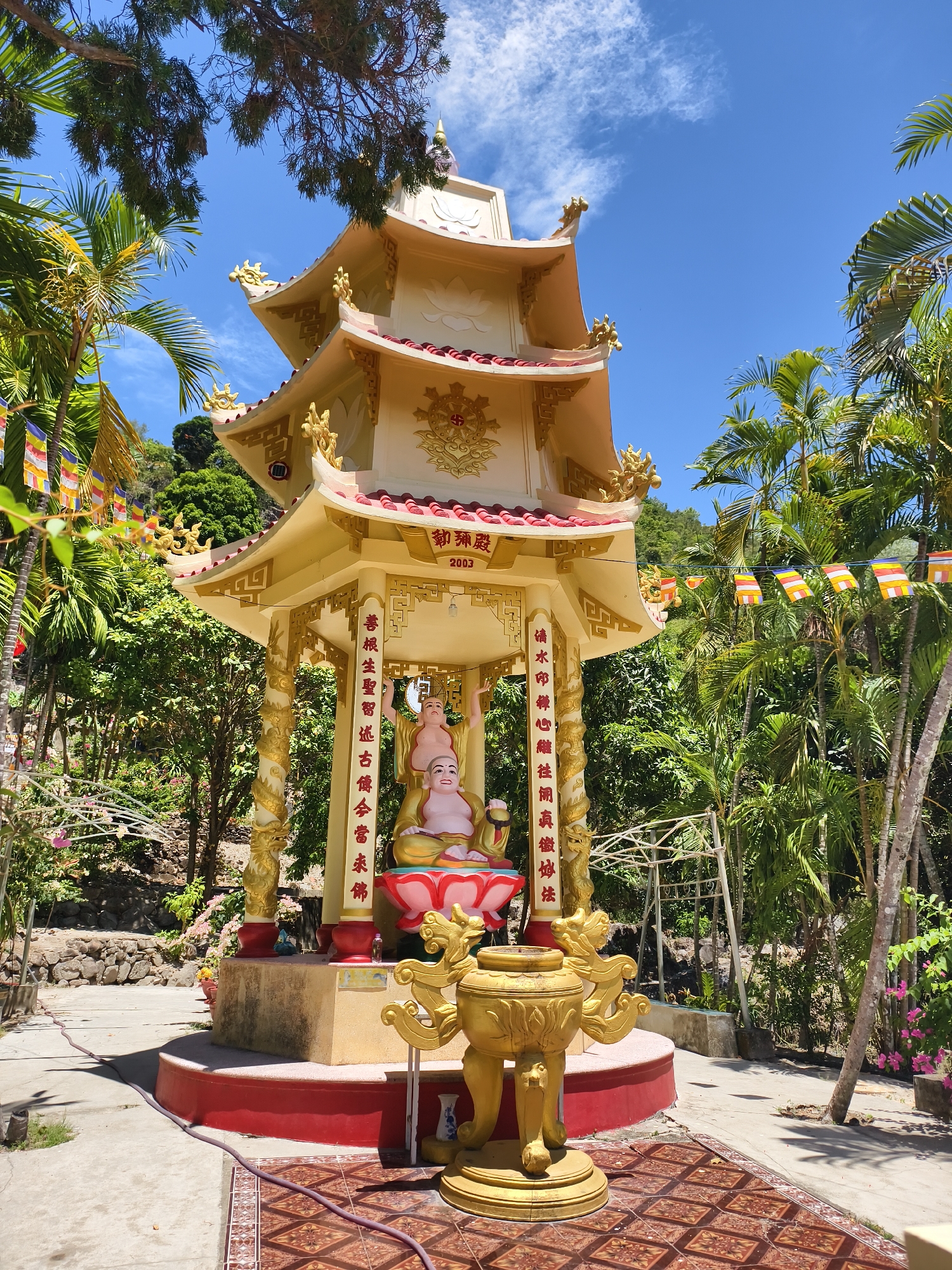 The image shows a three-tiered pagoda-style structure in Tay Nha Trang Ward, Vietnam. The pagoda is predominantly gold with red accents on the roofs and base. Within the pagoda, two statues of Buddha figures are seated on a pink lotus platform. A golden censer sits in front of the pagoda. The structure is surrounded by lush greenery, including palm trees and other tropical vegetation. Prayer flags are strung between the trees. The sky is blue with some wispy clouds. The pagoda has Chinese characters on the front, with the year "2003" also displayed.