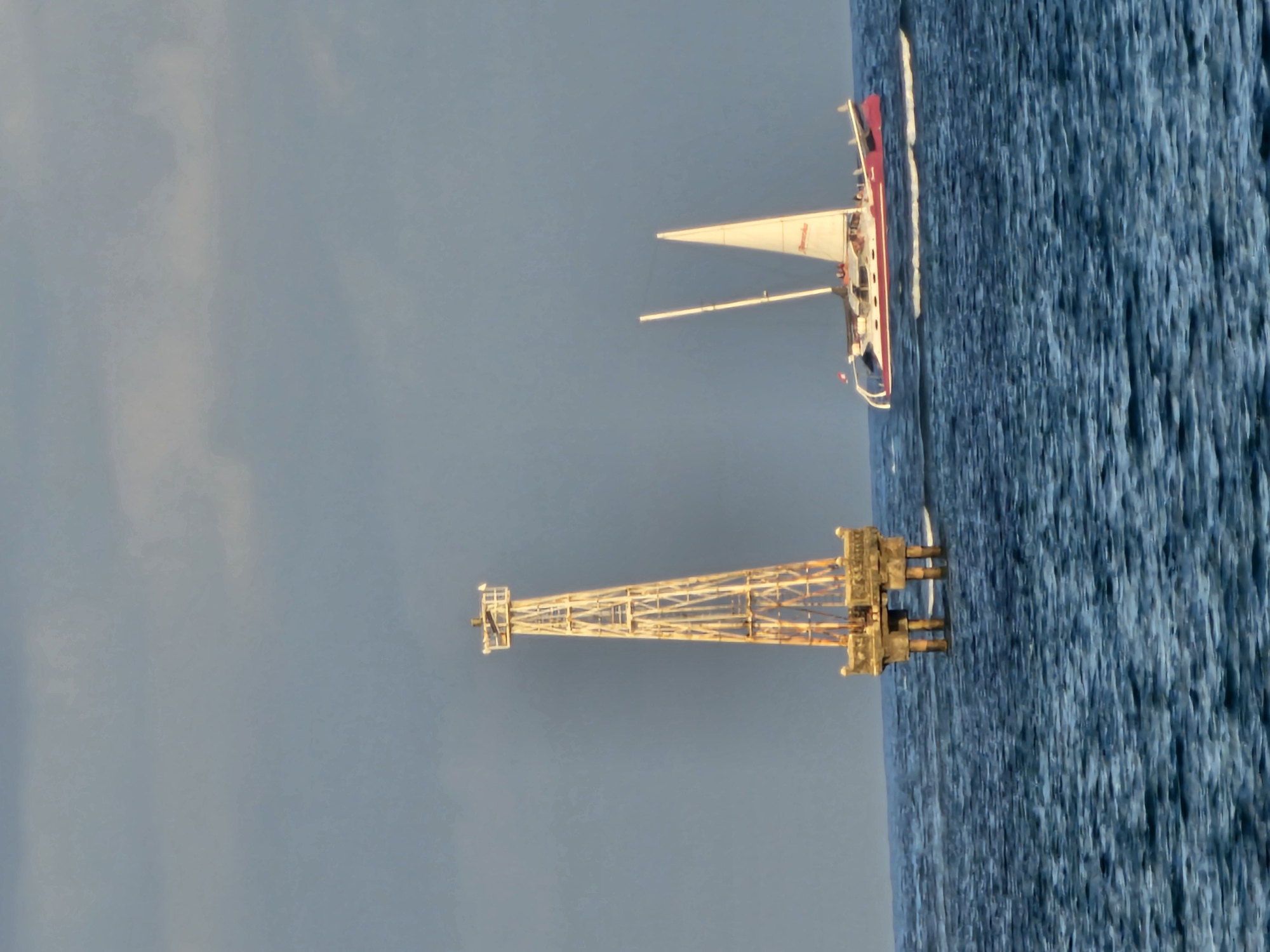 An offshore platform stands in choppy blue water under a hazy, overcast sky. The platform, a tall, white lattice structure with a square top section, is supported by four thick concrete pillars extending from a square base. To the right of the platform, a red and white sailboat with two masts and white sails is moving through the water. The sailboat has a small flag attached to its stern. The scene appears to be on the coast of Denpasar, Indonesia.