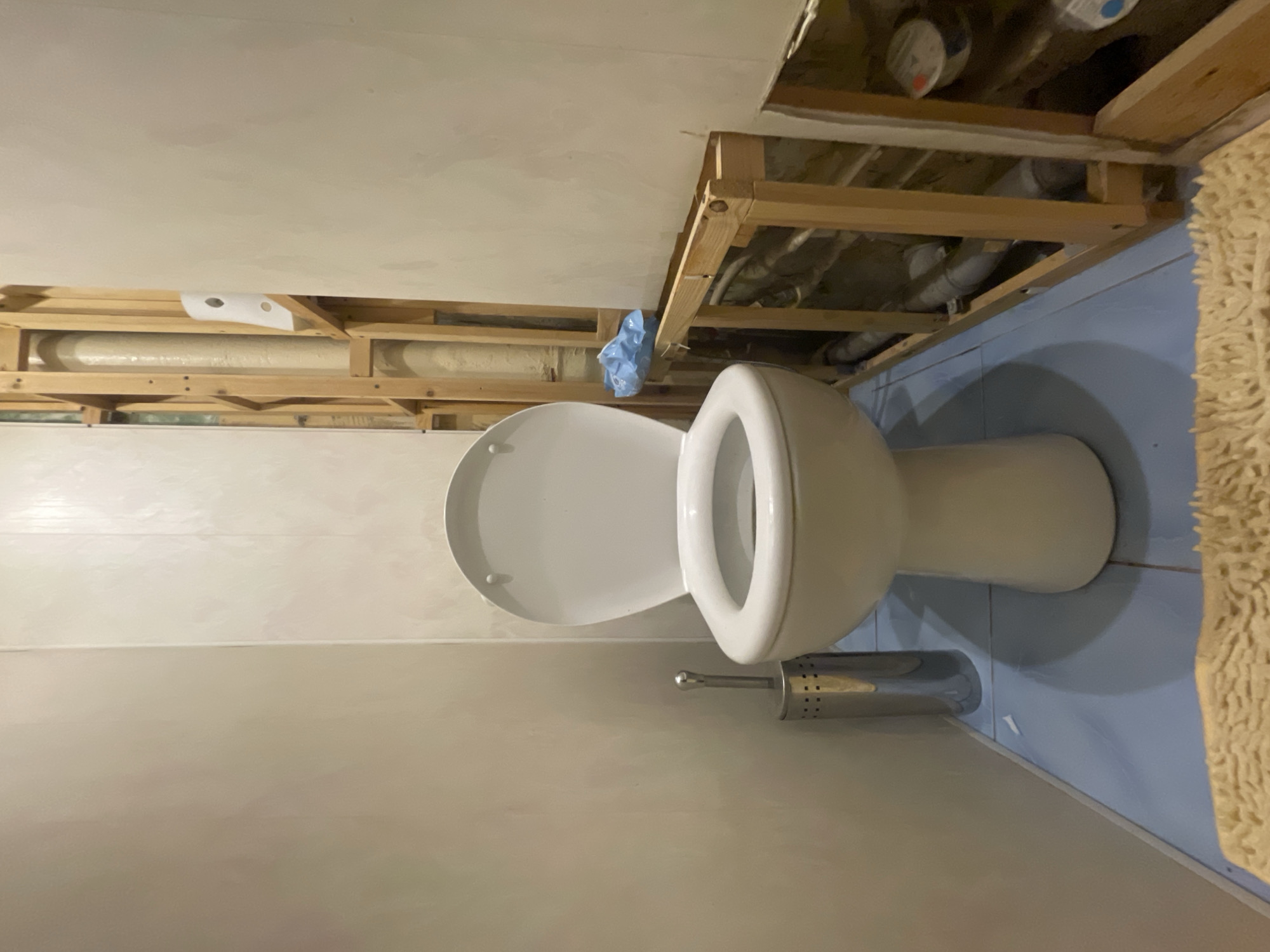 The image depicts an unfinished bathroom with a white toilet in the center. To the left of the toilet is a silver toilet brush holder. The walls are a light beige color, with the right wall exposing exposed wooden studs and plumbing. The floor is tiled in a light blue color. A tan rug is partially visible in the foreground. The overall scene suggests a renovation or construction in progress. The location in Yegoryevsk, Russia, is not visually discernible from the image content.