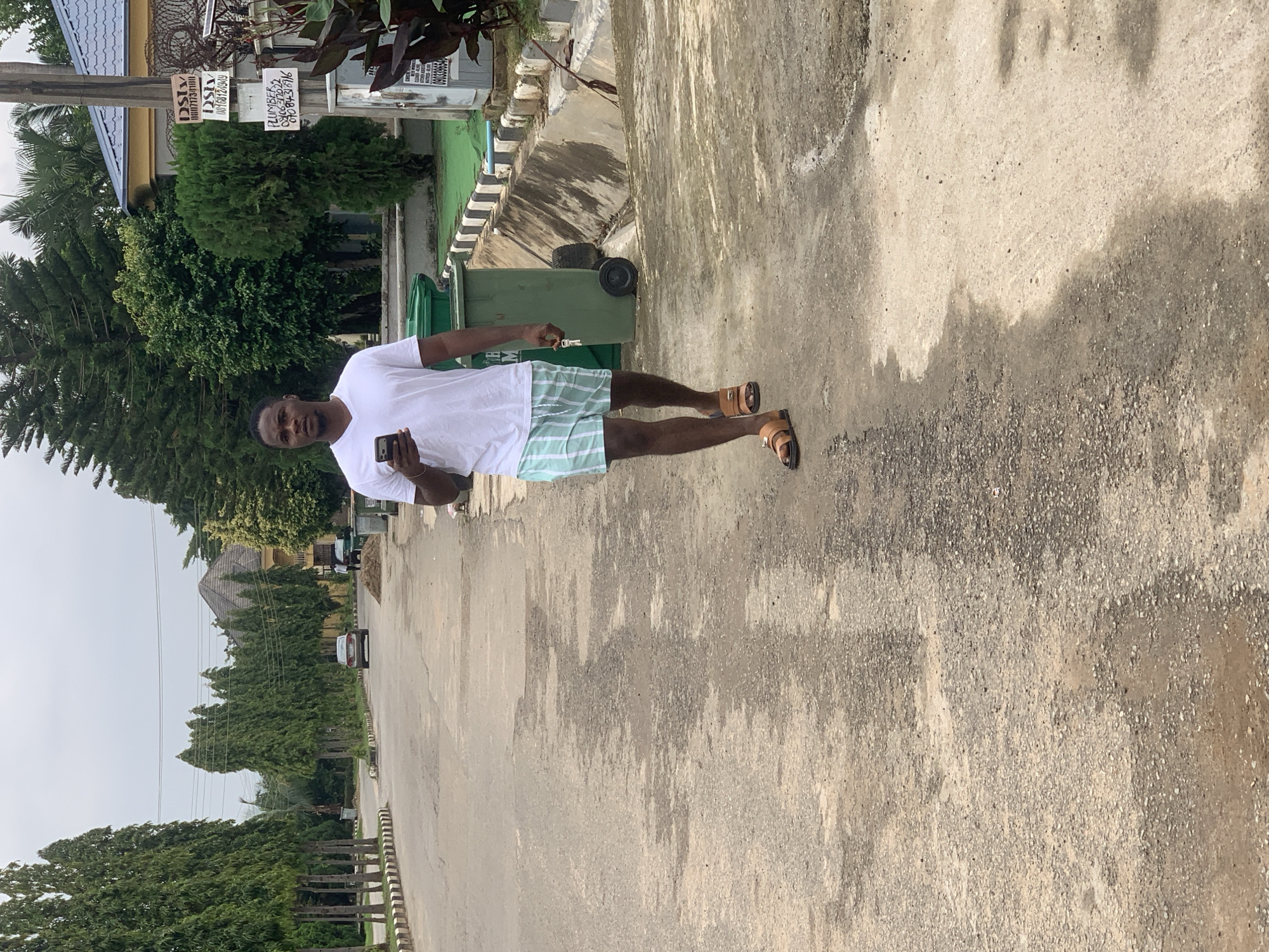 A young Black man is walking on a paved road in Abuja, Nigeria, holding a smartphone in his right hand and a set of keys in his left. He is wearing a white V-neck t-shirt and light blue and white striped shorts, with brown sandals on his feet. The road is lined with tall, manicured trees on both sides, and there are residential buildings and houses in the background. A green dumpster is visible on the right side of the road, and there is a car parked further down the street. The sky is overcast, suggesting a cloudy day.