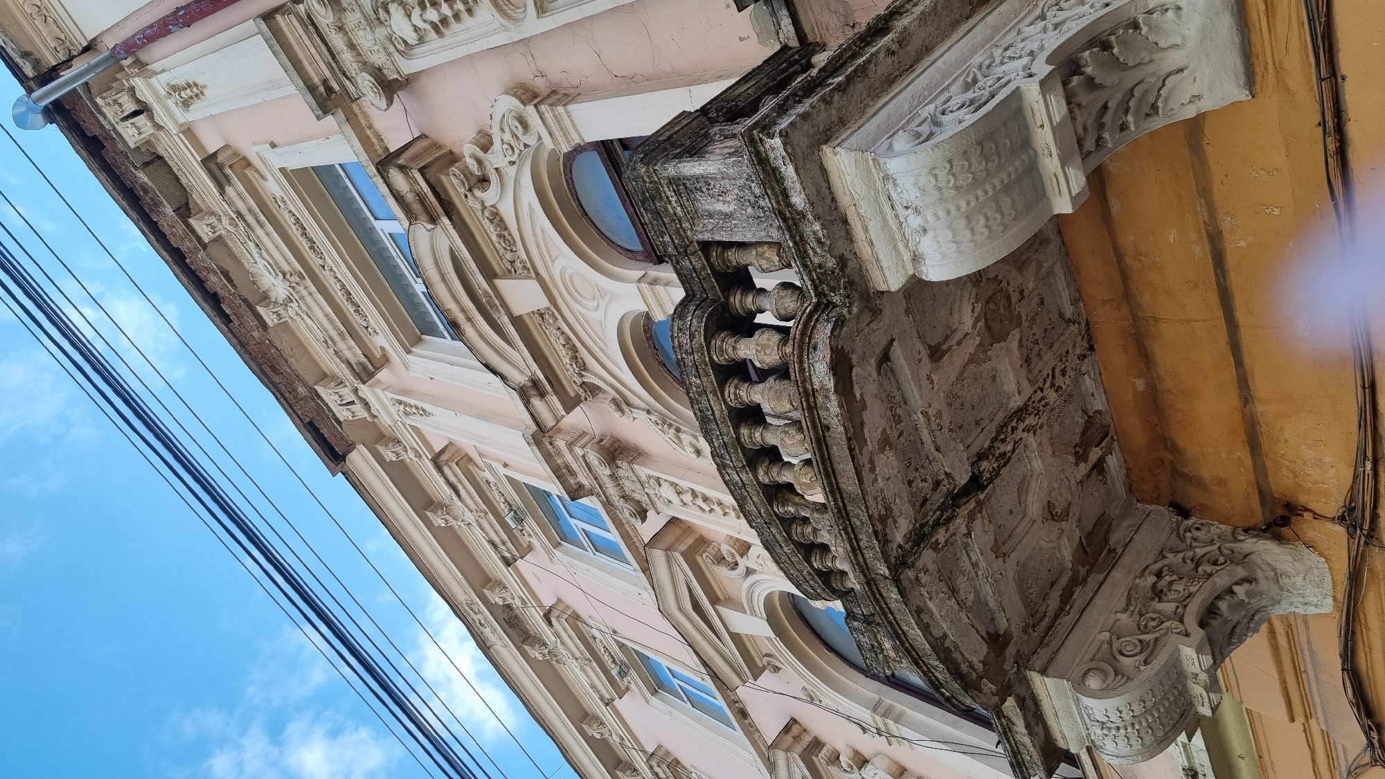 The image is an exterior, low-angle shot of a weathered, ornate balcony on a building in Chernivtsi, Ukraine. The building's facade is painted a light pink, with cream-colored decorative elements. The balcony itself is made of stone, with a balustrade of turned spindles and a cracked, peeling underside. Intricate carvings adorn the supporting brackets of the balcony. Above the balcony, windows with arched tops are visible, also framed by decorative molding. The building's roofline appears damaged, with exposed brickwork. Overhead, multiple black electrical wires are strung across the bright blue sky, which has a few wispy white clouds. The lower part of the building is a contrasting yellow color. The scene suggests an older, possibly historical building in a state of disrepair, juxtaposed against a clear sky and urban infrastructure.