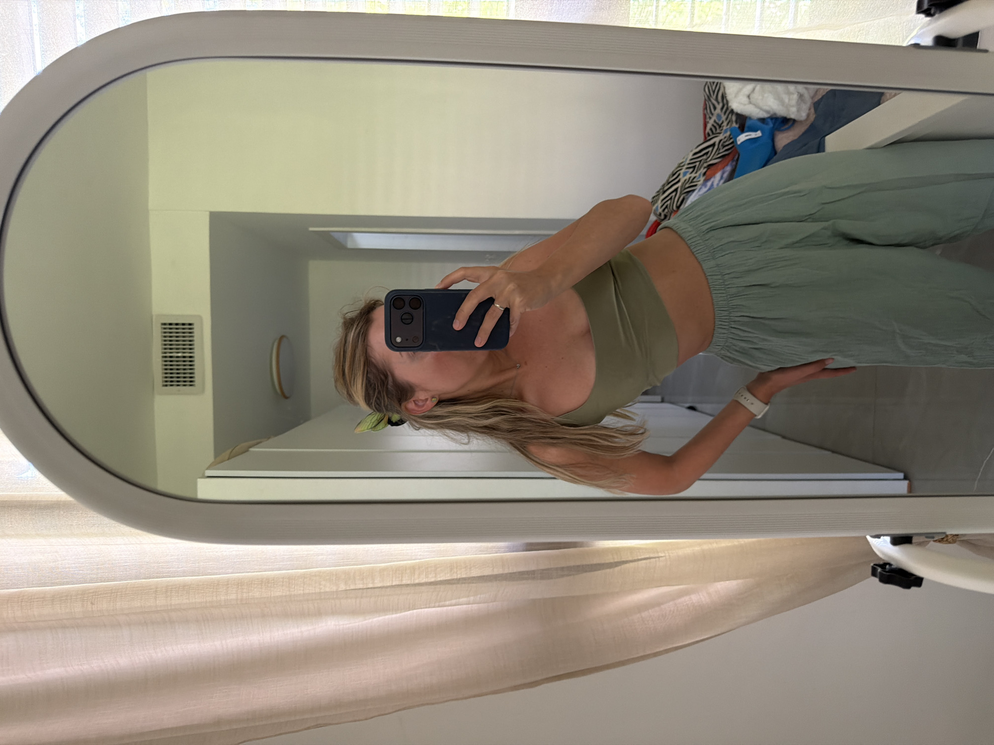 In a residential interior, a young woman with blonde hair is taking a mirror selfie. She is wearing a green crop top and light green wide-leg pants. A green butterfly clip is in her hair. Her right hand is holding a dark blue phone, and her left hand is resting on her hip. A silver ring is on her left ring finger. She is wearing a white smartwatch on her left wrist. The mirror reflects a hallway with white walls and a white ceiling vent. A light-colored curtain hangs on the left side of the frame. Clothes are piled on a white surface in the reflection to the right of the woman. The scene appears to be in Bat Yam, Israel.