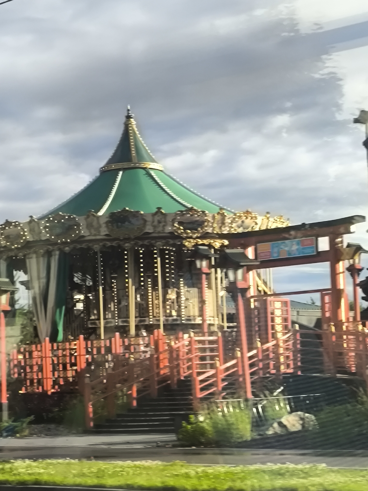 The image shows an outdoor carousel with a green, conical roof. The carousel is decorated with lights and ornate gold trim. A red, Japanese-style torii gate stands in front of the carousel, framing a sign with colorful illustrations. Red railings and stairs lead up to the entrance of the carousel. The scene appears to be a theme park or amusement area. The sky is cloudy, suggesting it might be a partly cloudy or overcast day. The lighting suggests it is daytime. The image is somewhat blurry and appears to have been taken from a moving vehicle. There is no discernible text on the sign other than the illustrations.
