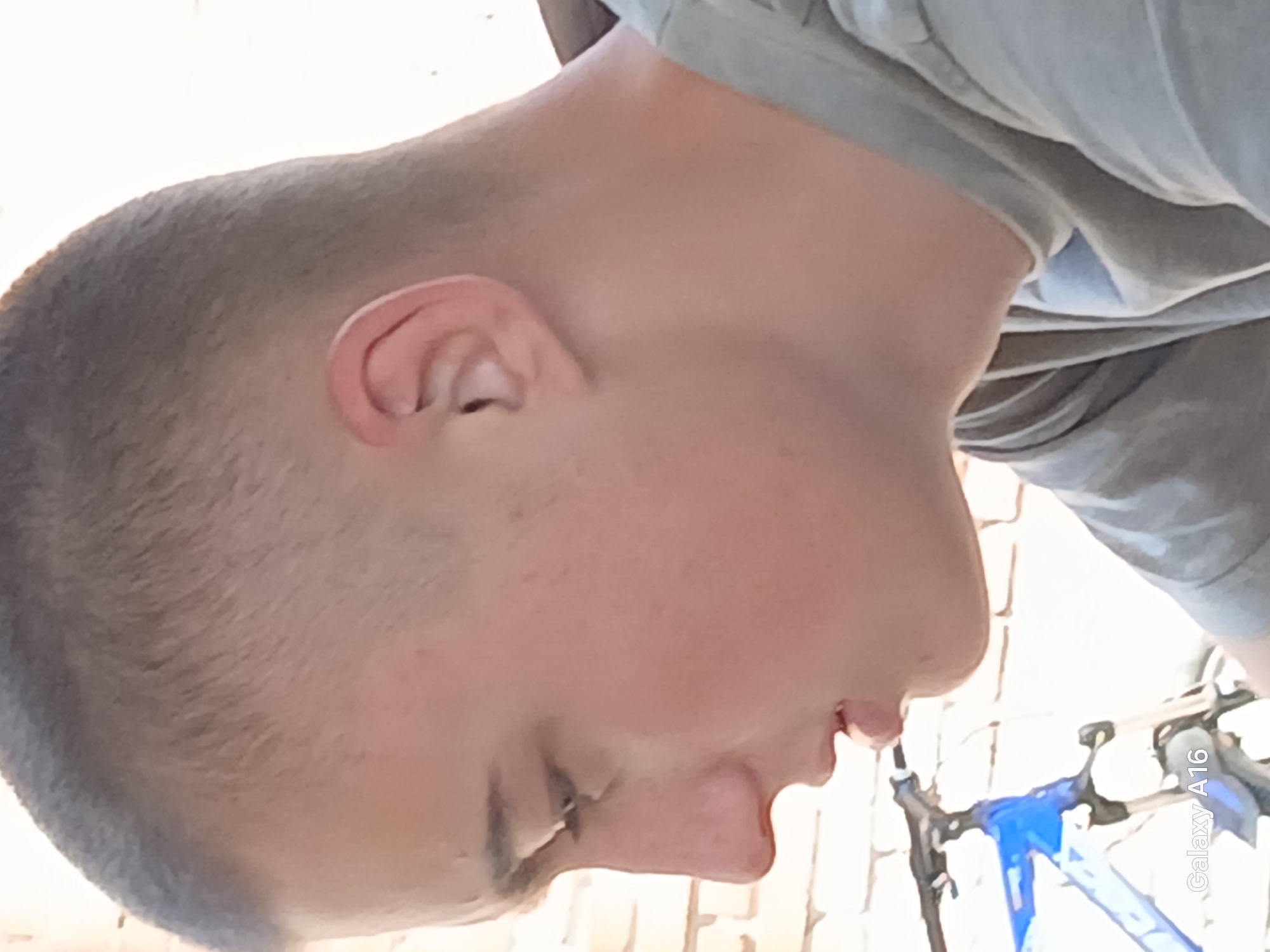 A young male, appearing to be in his late teens, is shown in profile, facing left. He has a closely cropped haircut and is wearing a grey t-shirt. The background consists of a brick wall and a blue bicycle with visible suspension forks. The lower left corner of the image displays the text "Galaxy A16". The image was captured in Ustyvytsia, Ukraine.