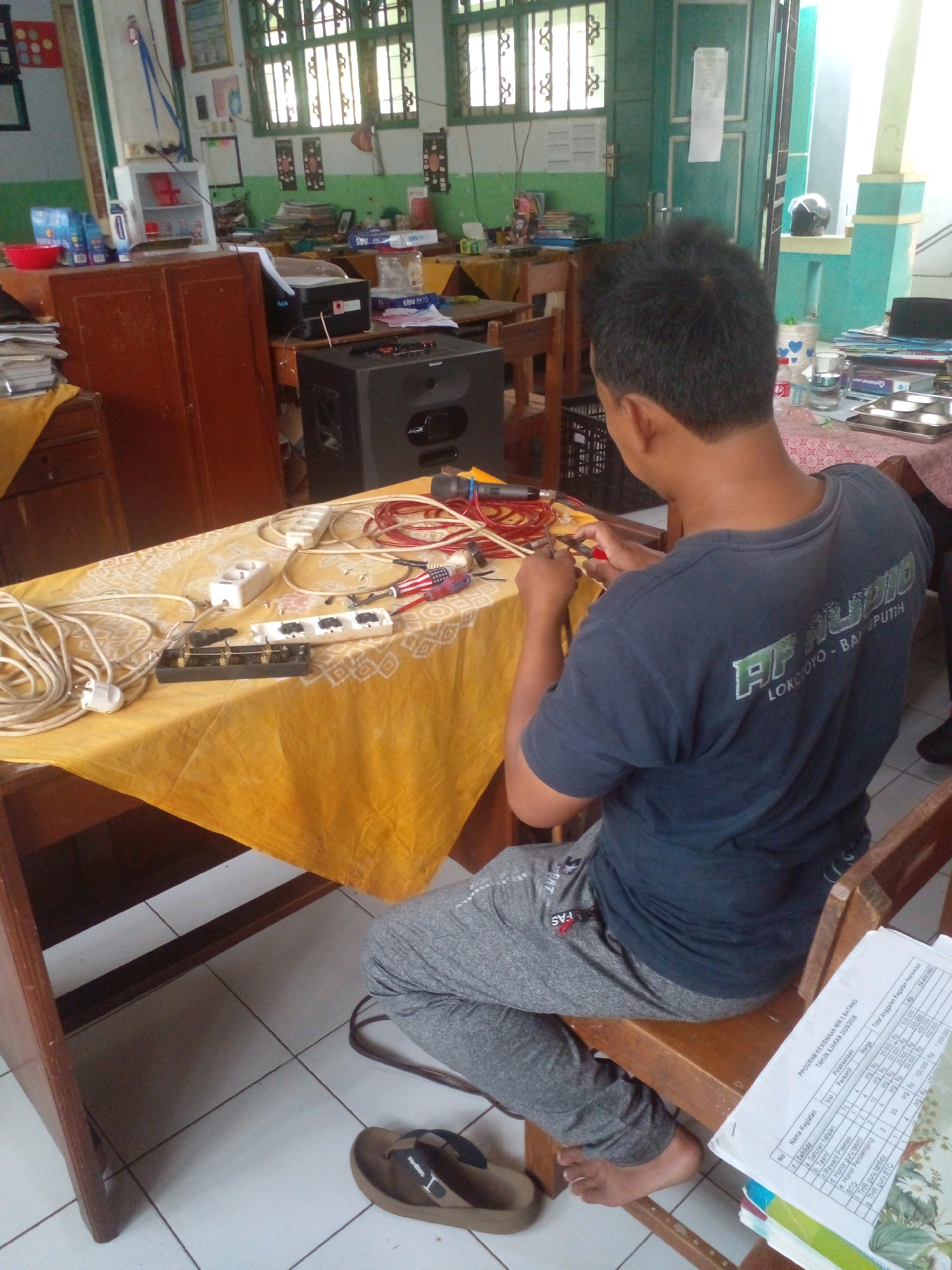 A man is seen from behind, wearing a dark blue t-shirt with a green logo and grey sweatpants, sitting on a wooden chair at a table covered with a yellow and white patterned cloth. The table is cluttered with electrical components, including extension cords, plugs, power strips, and what appears to be wiring tools. The man is holding a pair of red-handled pliers and seems to be working on the electrical setup. In the background, there is a dark speaker, a wooden cabinet, shelves filled with books and other items, and windows with green frames. The overall setting suggests a room that could be a workshop or a classroom in Banyuputih, Indonesia, given the text visible on the document to the right of the man, which includes "Banyuputih" as part of an address. The floor is tiled with white and grey squares.