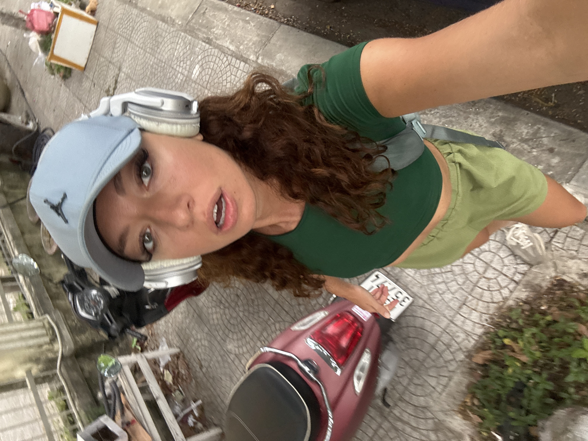 A woman with curly brown hair, wearing a green crop top, olive shorts, a light blue baseball cap, and silver headphones, takes a selfie with a maroon scooter on a patterned concrete sidewalk. The scooter has a license plate with the number 35774. Other objects in the background include a black motorcycle, potted plants, trash, and a building. The location is likely in Đà Nẵng, Vietnam.