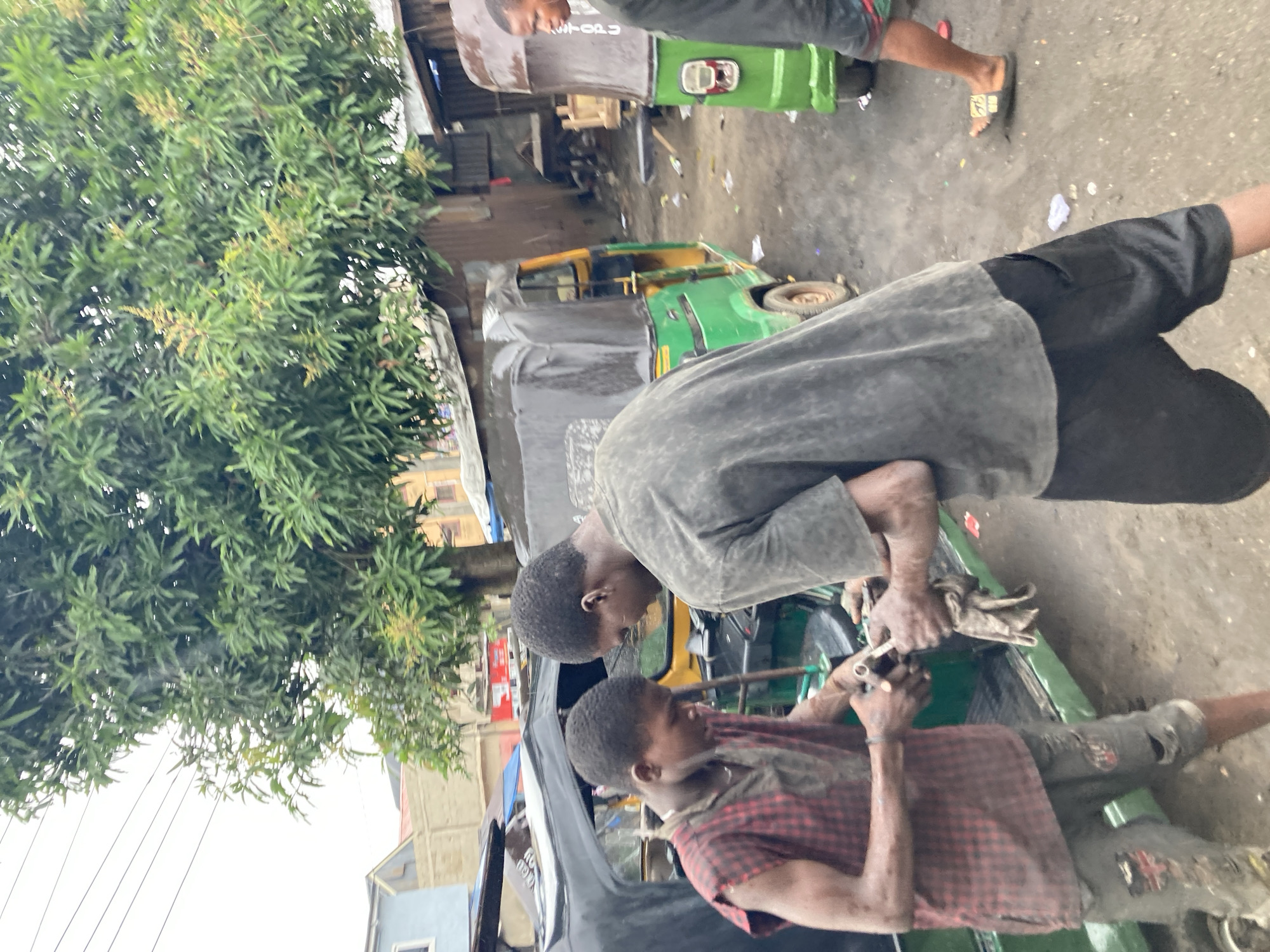 In Umuode, Nigeria, two individuals work on a motorized tricycle (keke). One person, wearing a grey shirt and black shorts, leans over the keke. The other person, wearing a red and black checkered shirt and soiled pants, holds tools. A third individual, wearing a grey t-shirt and green shorts, is visible in the background. The scene takes place in a cluttered roadside environment with buildings and a large tree in the vicinity. Another motorized tricycle is parked behind the one being repaired. The ground is soiled.