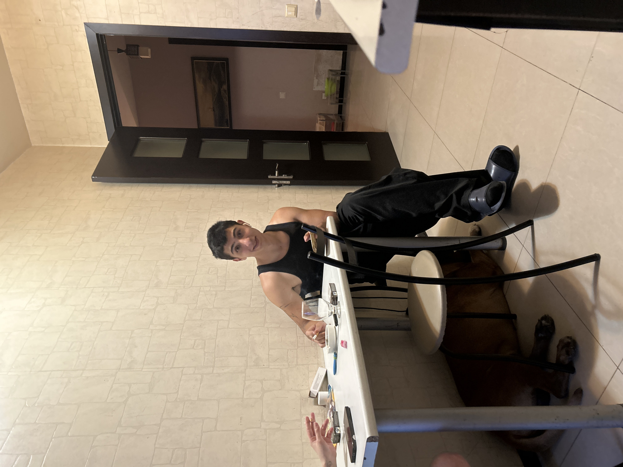 The image shows a person sitting at a white table in what appears to be a kitchen or dining area. The person is wearing a black tank top and black pants, with blue slide sandals. They are looking at the camera and smiling. On the table are various objects, including a wine glass, a cigarette, a phone, and some packaging. A dog is lying on the floor under the table. In the background, there is a doorway leading to another room, which is dimly lit and contains some shelves with items on them. The walls are tiled with a light-colored, stone-like pattern. The lighting suggests it is daytime, possibly indoors with artificial lighting. The overall mood seems relaxed and casual.