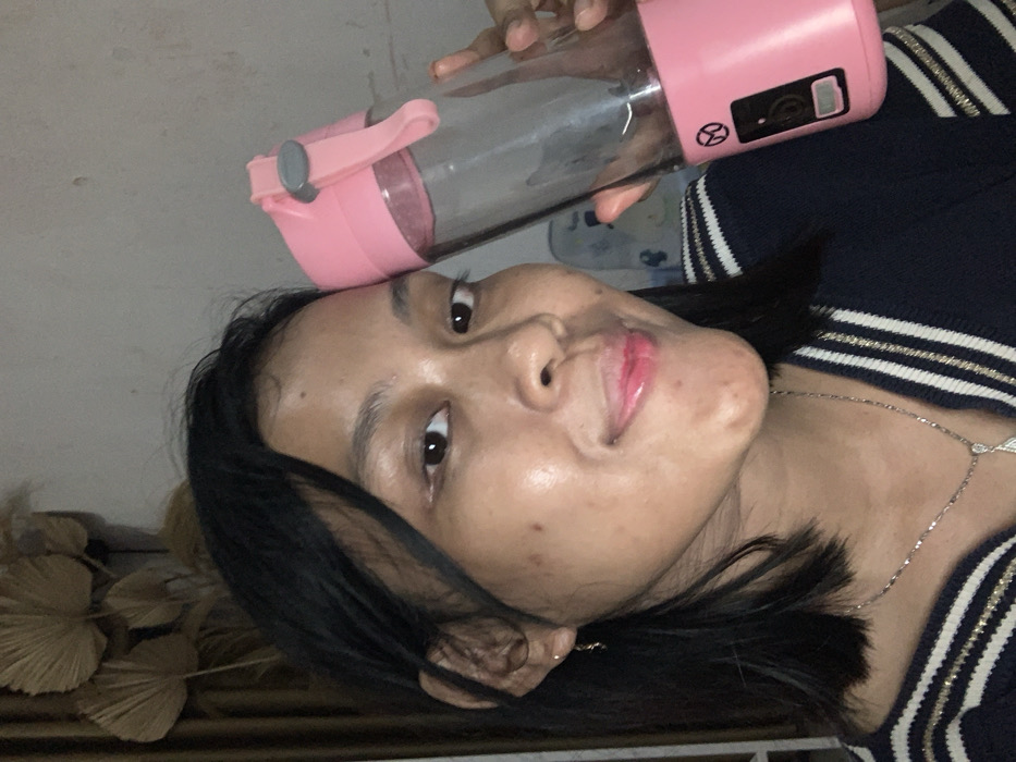 A female subject is depicted in a close-up, indoor setting within Kecamatan Pati, Indonesia, capturing a self-portrait. She possesses dark, shoulder-length hair, styled with a side part and some strands framing her face. Her complexion shows minor blemishes and a mole on her left cheek. She is wearing small gold-colored hoop earrings and a silver-toned necklace with a pendant. Her attire consists of a dark-colored top or sweater featuring horizontal white and gold stripes on the collar/shoulder area. Her expression is a slight smile, with her gaze directed towards the camera. She is holding a clear portable blender with pink top and base sections in her right hand, positioned beside her head. The blender features a grey carrying loop on its lid and a control panel on its base, including a circular symbol with a diagonal line. The background reveals a light-colored wall adorned with several dried, fan-shaped leaves arranged decoratively, and a brown curtain or fabric partially visible on the left. The lighting is subdued.