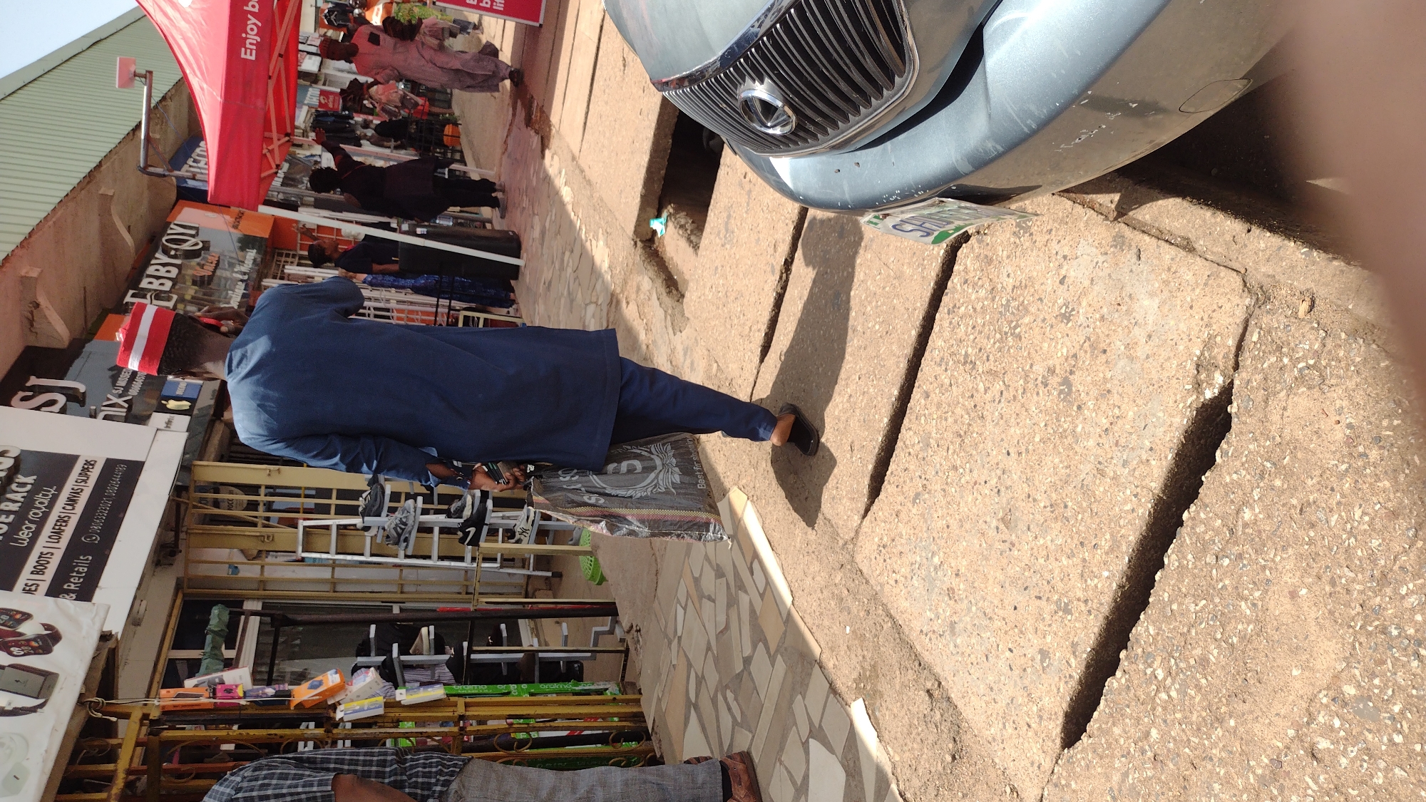 The scene captures a street in Jos, Nigeria. A man in a blue traditional attire, complemented with a red-and-white striped cap, is walking on a paved sidewalk. He carries a dark-colored shopping bag in his left hand. The sidewalk is adjacent to a storefront, marked by a yellow support structure laden with items for sale, including shoes displayed on shelves. The shop sign reads "SJ...WEAR ROYALTY...RETAILS." Across the street, vendors and customers are gathered under a red awning. A light-blue vehicle is parked on the edge of the sidewalk. The ground around the vehicle shows a gap in the paving.