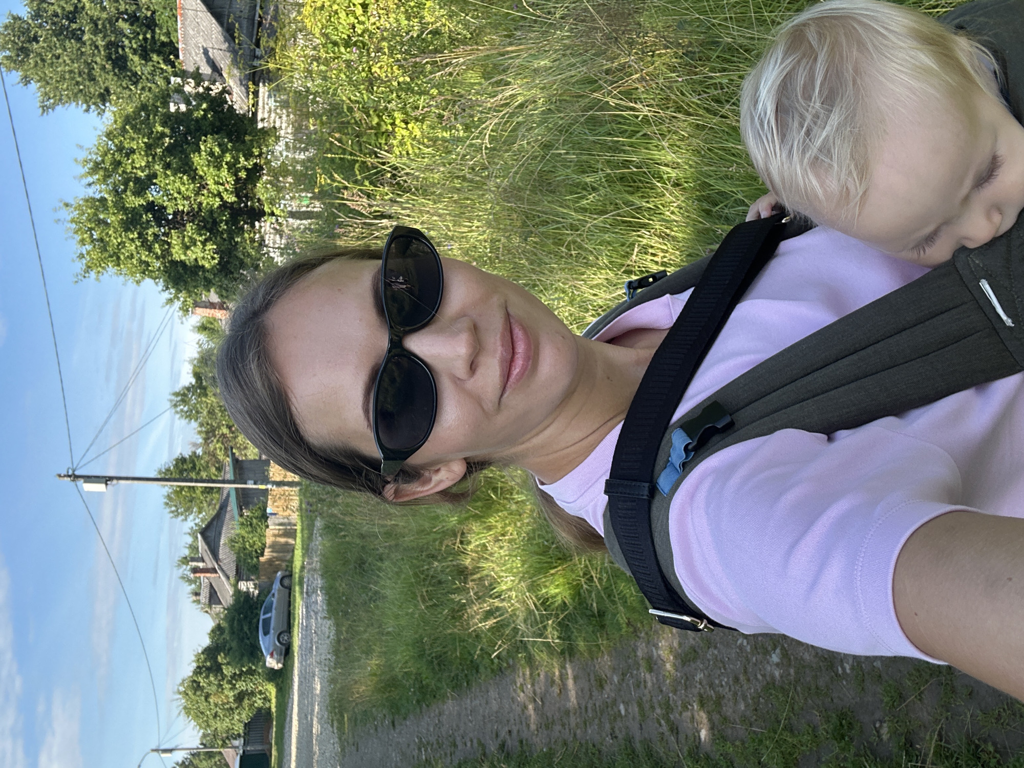 In Slantsy, Russia, on a dirt path beside a grassy area with tall weeds, a woman is taking a selfie. She is wearing a light pink shirt and black sunglasses. Her hair is pulled back, and she is smiling. She is carrying a baby in a dark green baby carrier on her chest. The baby, with blonde hair, is asleep. In the background, there are houses with grey roofs, a silver car, and trees. The sky is blue with some clouds.