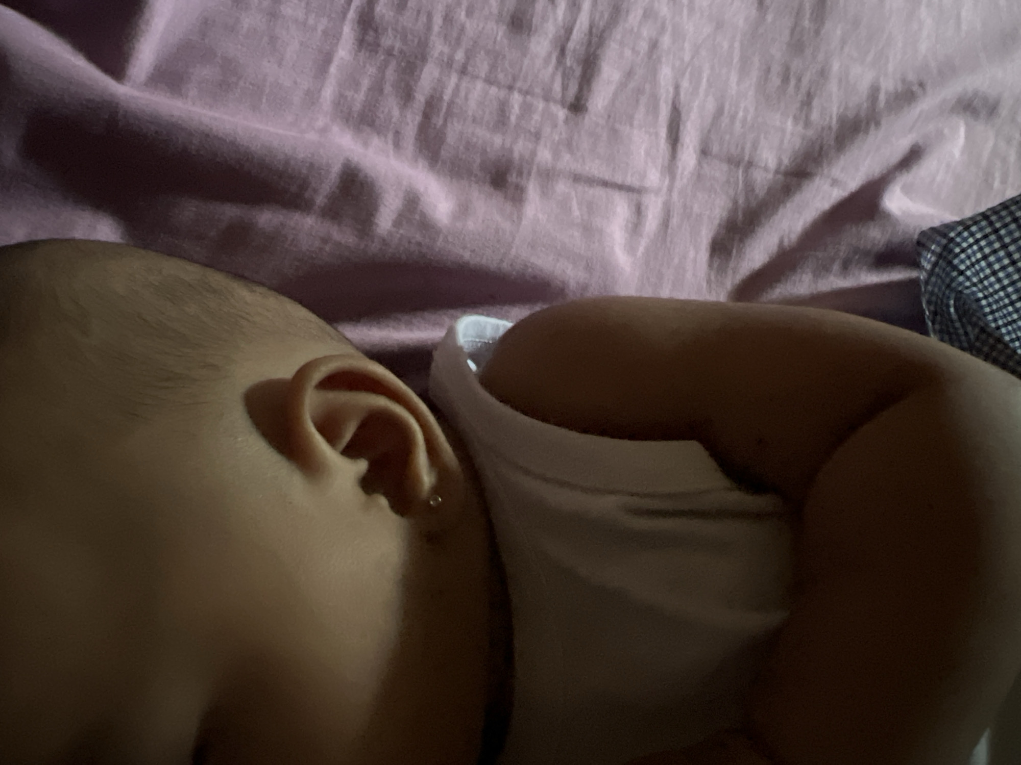The image shows a close-up, overhead view of a baby's ear and shoulder, lying on a pink textured surface. The baby has fair skin and a small silver earring in its left earlobe. The baby is wearing a white sleeveless top, and a plaid fabric is visible at the bottom edge of the frame. The lighting suggests an indoor setting, likely a bedroom. There is no discernible connection to Lhokseumawe, Indonesia, as the image provides no geographical context.