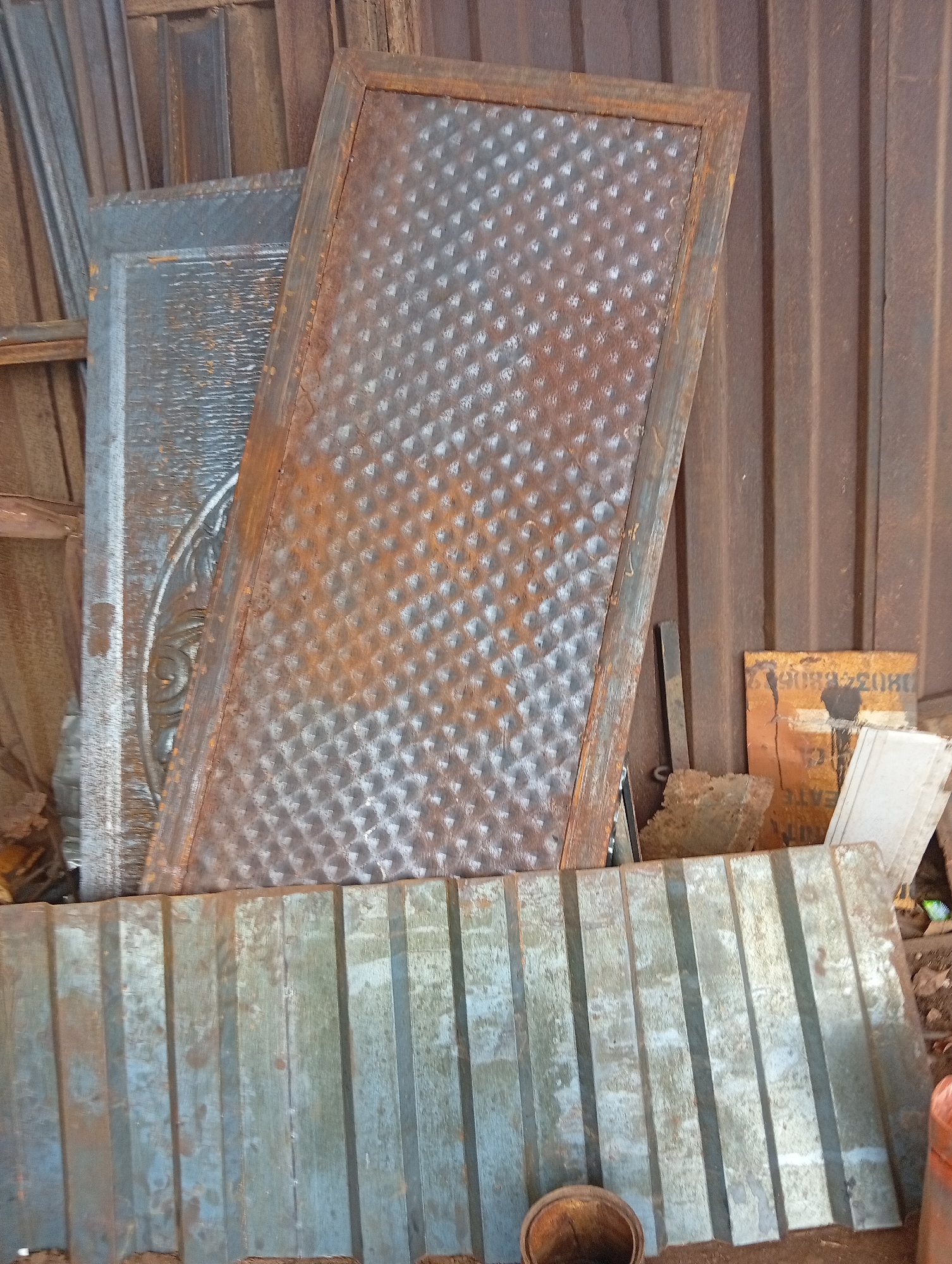 The image depicts a collection of weathered metal materials, suggesting a storage area, scrap yard, or workshop setting in Owerri, Nigeria. The scene is brightly lit, indicating daytime.
In the foreground, a large, horizontal sheet of corrugated metal lies on the ground. It appears worn, with patches of light blue-grey paint or coating mixed with areas of rust and dirt. Below this sheet, a small, rusty, cylindrical metal container or bucket is partially visible.
Behind the corrugated sheet, several other metal panels are propped up. The most prominent is a tall, rectangular panel positioned at an angle, displaying a repeating raised diamond pattern across its surface. This panel is heavily rusted, exhibiting shades of orange, brown, and mottled grey. To its left, a second, wider metal panel, mostly greyish, is partially visible behind the diamond-patterned one. A section of this panel reveals an elaborate, possibly embossed or cast, circular decorative motif. Further left, other narrow metal pieces or beams lean against the background.
The background consists of a vertical corrugated metal structure, likely a wall or fence, also showing signs of rust and weathering. To the right of the central panels, a piece of cardboard or similar material with an orange-brown hue is propped up. Visible text on this material includes a sequence of numbers starting with "0803" and the partial word "EATE". Small, irregular pieces of rusty metal scrap are scattered on the ground around the base of the propped materials. The overall impression is one of disused or awaiting-reuse industrial materials.