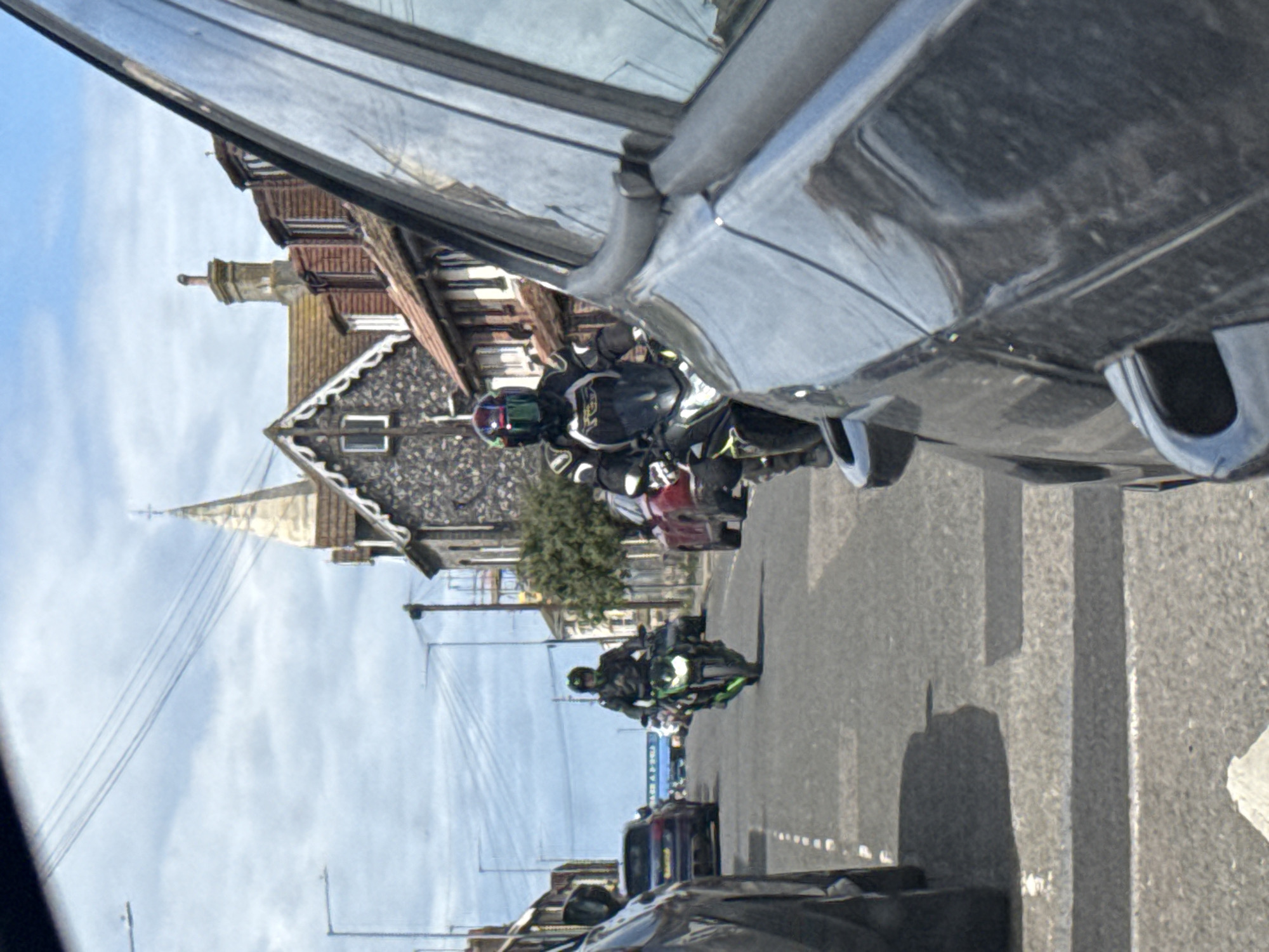 The image, shot from inside a vehicle, captures a street scene in Great Yarmouth, United Kingdom. A convoy of motorcyclists rides along a road lined with buildings on either side. The motorcycles are varied in color, with green and red examples visible. The road is marked with painted lane dividers. A variety of other vehicles are also visible on the road. Buildings of brick, stone, and tile feature in the background, exhibiting details such as pointed roofs, chimneys, and decorative gables. Overhead, utility lines traverse the sky, which is lightly clouded. The interior of the car in which the image was taken is a prominent element of the foreground, partially obscuring the view.