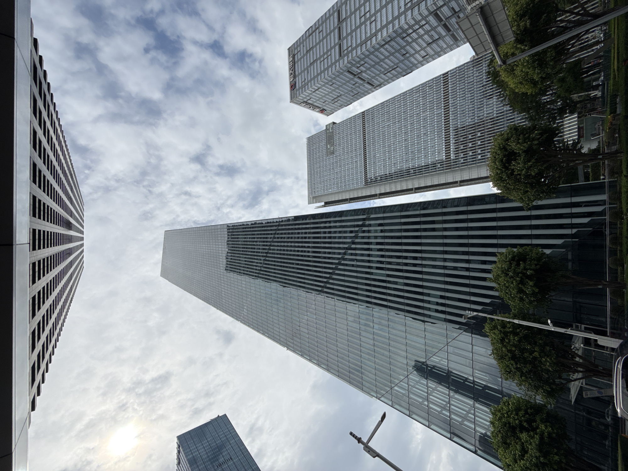 This is a wide-angle, low-angle photograph of a cluster of modern skyscrapers in Tianhe District, China, on a cloudy day. The sun is visible through the clouds on the left side of the frame, casting a slight glare. The buildings are tall and appear to be made of glass and steel, with a variety of architectural styles, including a prominent building with a reflective, angled glass facade and another with a grid-like pattern of windows. The foreground shows the tops of several green trees, suggesting a street-level view looking upwards. A lamppost with a modern design is visible on the left. The overall impression is one of a bustling, contemporary urban environment.