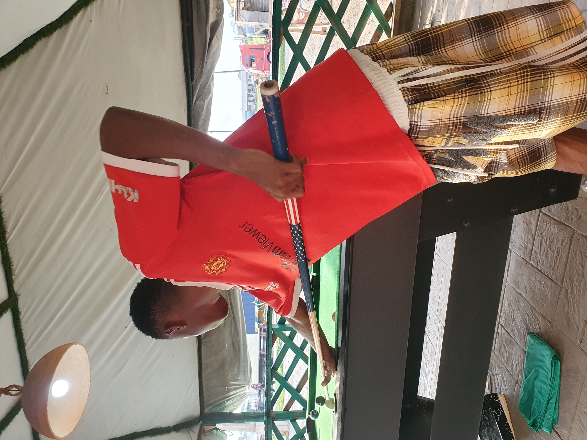 In Lokoja, Nigeria, a young Black male is pictured playing pool. He is wearing a red Manchester United jersey, a white headband, and plaid shorts. The pool table is green, and there are several balls on it, including a cue ball, a solid blue ball, and a striped yellow ball. A cue stick, decorated with an American flag pattern, is positioned to strike the cue ball. The scene takes place outdoors, under a thatched roof, with a fence and some trees visible in the background. A truck is also visible in the distance. The ground is paved with stone tiles.