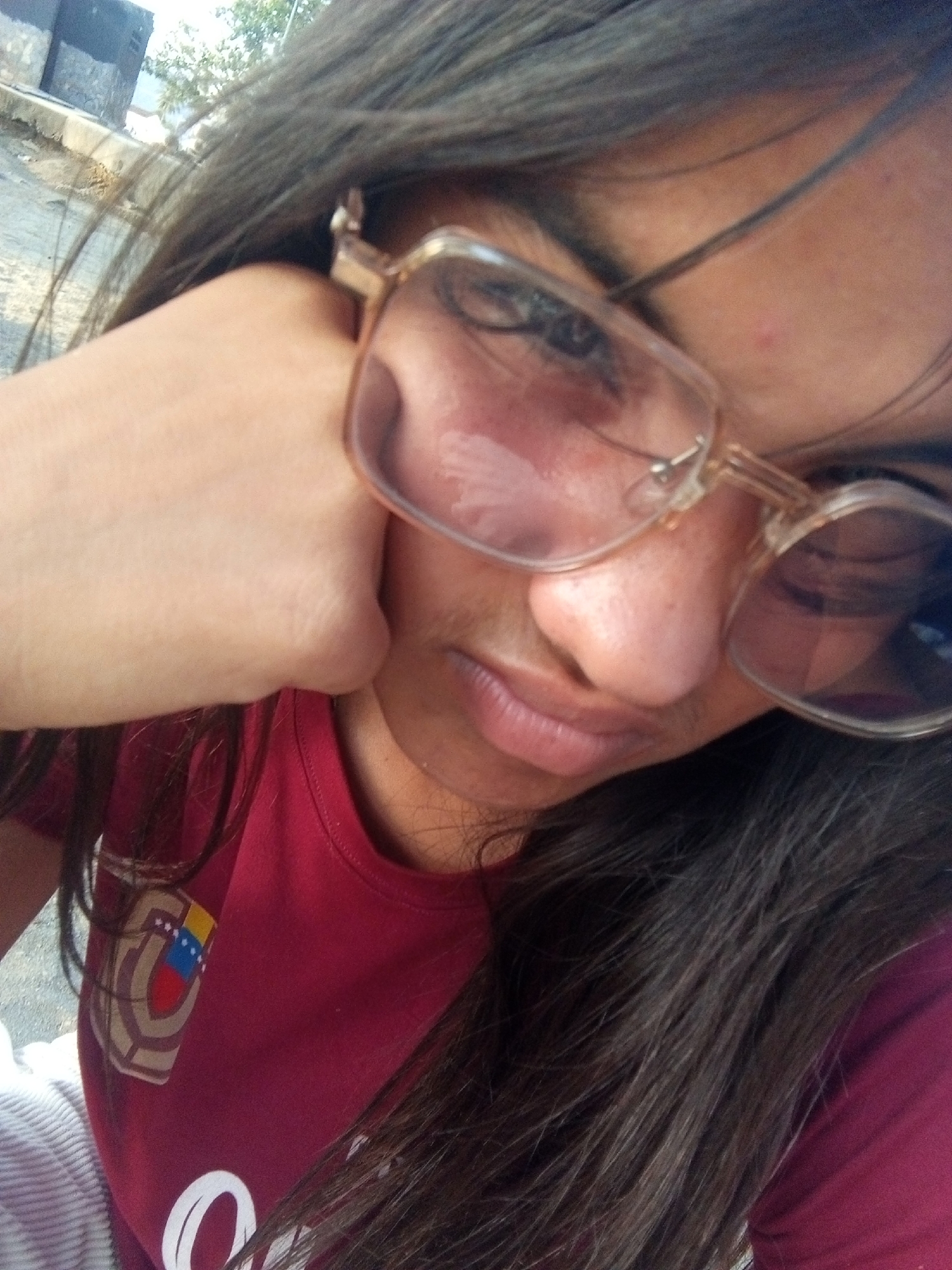 A close-up selfie depicts a woman with long, dark hair, resting her chin on her hand. She wears eyeglasses with a transparent frame. Her shirt is maroon and features a patch that includes the colors of the Venezuelan flag. The background is somewhat blurry, revealing some hints of an outdoor environment, possibly a street or paved area with some greenery. The location cannot be determined to be Juan Griego, Venezuela from the available visual information.