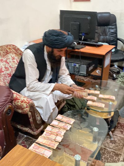 A man with a dark beard and a dark turban sits on a patterned armchair in a room. He wears a white tunic and a dark vest. He is counting stacks of currency on a glass-topped coffee table. A computer monitor, keyboard, and tower sit on a small desk behind the table. An office chair is behind the desk.