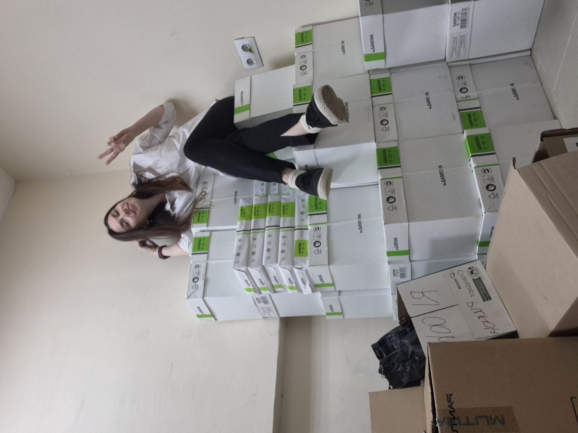 The image shows a person posing on a stack of boxes. The person, wearing a white shirt and dark pants, is lying on the boxes with their legs extended and feet pointed upwards. One hand is raised in a peace sign. The boxes are white with green accents and appear to be labeled with a name or logo. The boxes are stacked against a wall with an electrical outlet visible. Several other boxes and a black plastic bag are in the foreground. The location appears to be an indoor storage or stockroom.