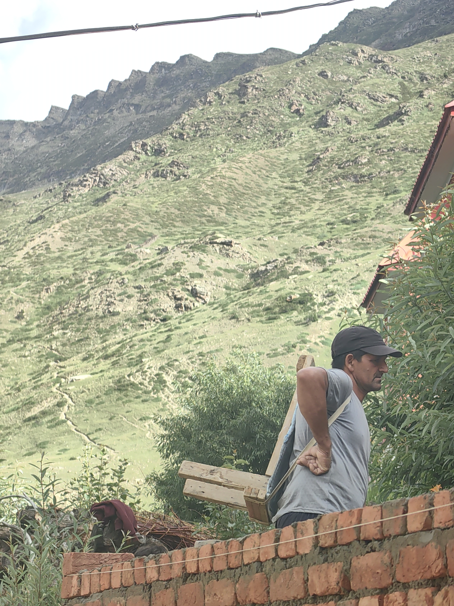 The media file shows a man in Keylang, India, carrying a wooden contraption on his back. He is wearing a grey t-shirt, a black baseball cap, and dark shorts. The wooden object appears to be a type of carrier or frame, possibly for transporting goods. Straps made of light-colored material are slung over his shoulders, supporting the load.
The man is seen from the side, in profile, looking towards the right of the frame. His right hand is resting on his lower back, a gesture that could indicate exertion or discomfort from carrying the weight. His expression is neutral.
The background depicts a mountainous landscape, characteristic of a hilly or high-altitude region. The mountains are covered in lush green vegetation, with patches of rocky terrain visible. A winding path can be seen ascending one of the slopes. In the foreground, a red brick wall runs horizontally across the lower part of the image. Behind the man and to the right, the roof and upper section of a building with a red roof are visible, partially obscured by dense green foliage. The overall lighting suggests it is daytime, with a slightly overcast sky. The scene conveys a sense of everyday labor in a rural, mountainous environment.