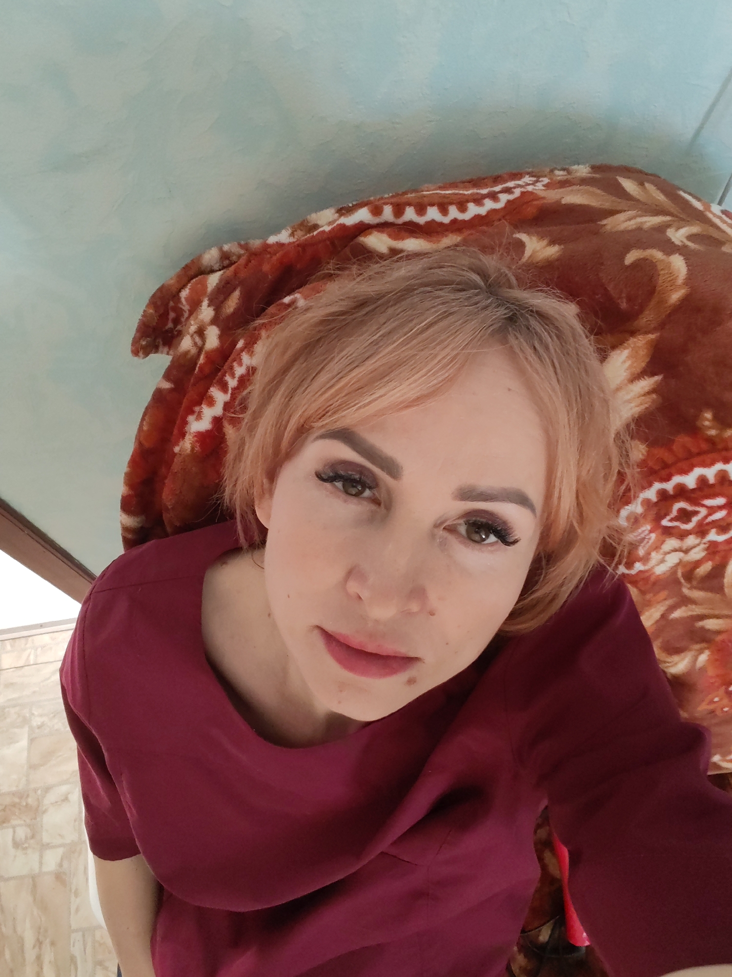 A medium-angle, close-up photograph shows a woman looking upward at the camera. She has light skin, auburn hair, and dark eyebrows. She is wearing false eyelashes, and her lips are red. She is wearing a maroon shirt. The background consists of a brown blanket with a floral pattern and a pale blue ceiling. The photo appears to be taken indoors. The location is unknown and cannot be inferred from the image.