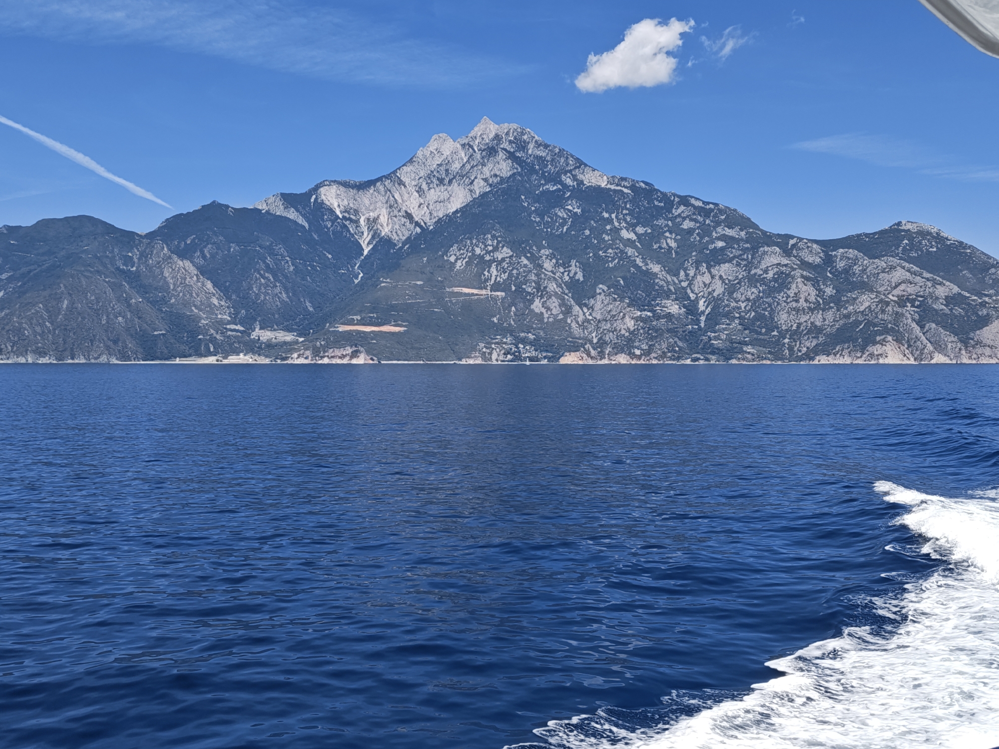 The image shows a mountain range with snow-capped peaks rising from the sea under a clear blue sky with a single white cloud. The sea is a deep blue, with white foam from the wake of a boat visible in the foreground on the right side of the frame. The mountains are rugged and appear to be covered in vegetation in the lower slopes, transitioning to rocky, snow-dusted peaks at higher elevations. The overall scene suggests a coastal view of a mountainous region in Greece, likely a prominent natural landmark visible from the sea.