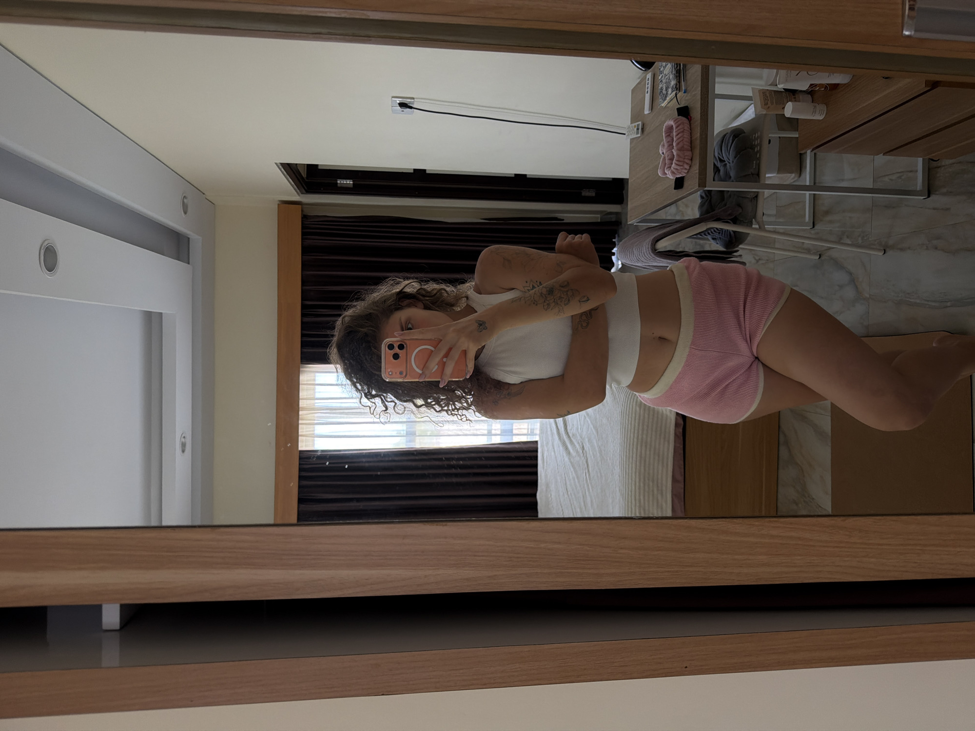 A young woman with curly brown hair, wearing a white crop top and pink shorts, stands in front of a full-length mirror in a room. She is holding a smartphone to take a selfie. Her arms are crossed. The room appears to be a bedroom or living space with a bed, a desk, and a wardrobe visible in the reflection. The walls are painted a light color, and there are dark curtains behind the bed. The flooring is tiled. The image is captured from a medium shot, focusing on the woman and her immediate surroundings. The scene is well-lit, likely by natural light coming from a window. The overall impression is that of a casual, personal moment captured in an indoor setting.