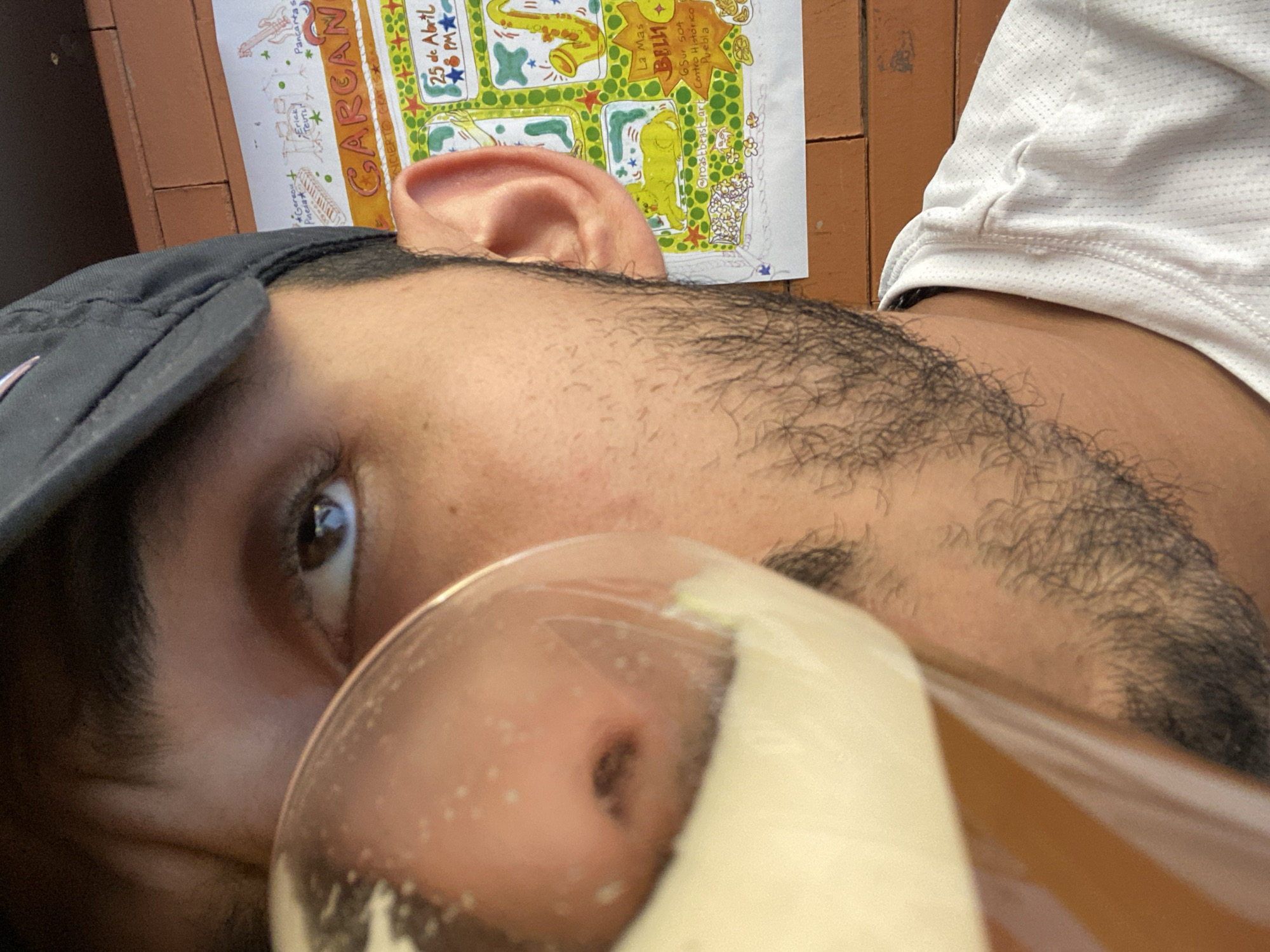 An eye-level close-up shot depicts a person holding a glass of liquid, possibly a beverage, with their face partially obscured by the glass. The person, with a baseball cap and facial hair, is wearing a white shirt. In the background, there is a poster with the word "GARGAN" visible, alongside the date "25 de Abril" and "8PM". The poster also includes "La Mas BELLI", "6Sur 504, Centro Historico, Puebla" and the Instagram username "@roastbeast_art". The setting appears to be in Puebla, Mexico, specifically within the Centro Histórico.