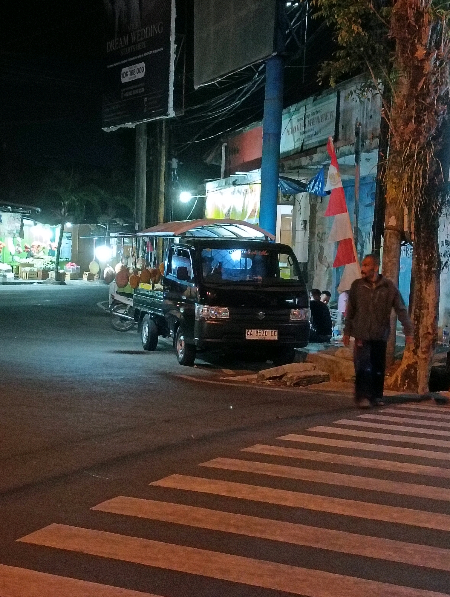 The image captures a nighttime scene on a street in Purworejo, Indonesia, featuring a black pickup truck parked near a pedestrian crossing. The truck is loaded with what appear to be durian fruits, hanging from its sides. In the foreground, a man is walking across the zebra crossing, his figure blurred due to motion. Several other people are visible in the background, some sitting near the truck. A street vendor's stall with bright lighting is on the left, displaying various items, possibly fruits or vegetables. Overhead, a large banner advertising "YOUR DREAM WEDDING STARTS HERE" is visible, along with numerous electrical wires. Red and white flags are also displayed, indicating a nationalistic or celebratory context. The street is paved, and the lighting suggests it is an urban area.