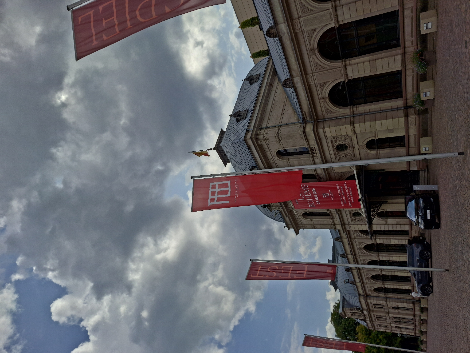 The image shows the exterior of the Festspielhaus Baden-Baden, a prominent opera house and concert hall located in Baden-Baden, Germany. The building, constructed from a light-colored stone, features a distinctive domed roof and arched windows. Red banners with white lettering hang from flagpoles in front of the building. One banner prominently displays the text "LA BOHÈME" along with a date, "23 AUG 2026". Another banner features a stylized window design and the text "FESTSPIELHAUS BADEN-BADEN". The sky above is cloudy, with patches of blue visible. Two cars, a dark SUV and a black sedan, are parked in front of the building's entrance. A single person in a light-colored outfit is walking near the cars. The ground is paved with cobblestones. The overall scene depicts the exterior of a cultural institution, likely before or after a performance.