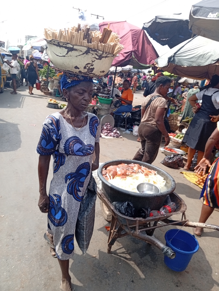An elderly woman balances a large bowl filled with wooden sticks on her head, walking on a busy street market. She's wearing a patterned dress and carrying a black bag. A rusty wheelbarrow beside her holds a large container overflowing with white and reddish food items, and a small blue bucket hangs from the handle. The marketplace is bustling with people and covered with colorful vendor umbrellas.