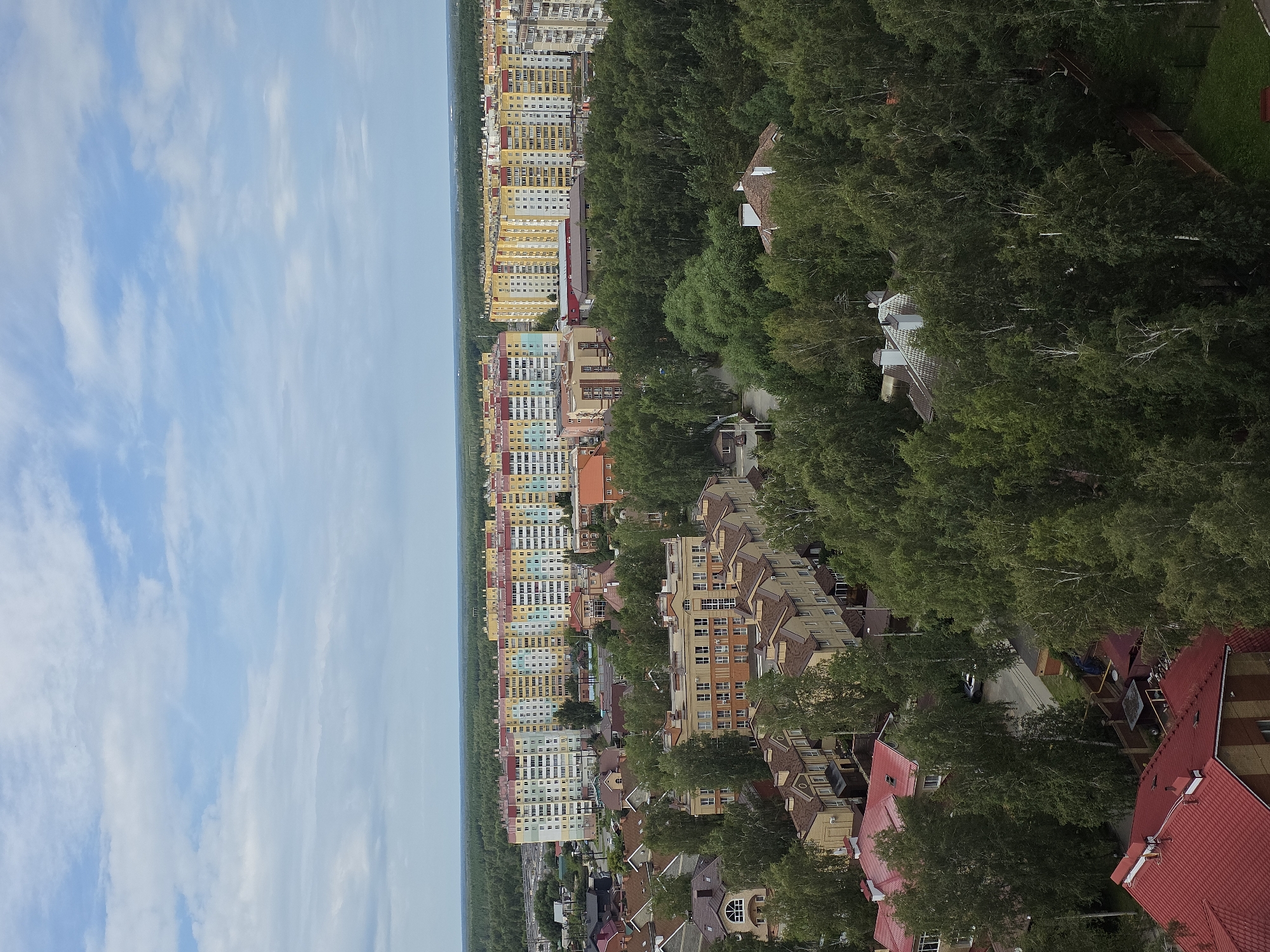 The image captures an aerial view of a residential area in Chelyabinsk, Russia, characterized by a mix of apartment buildings and smaller houses nestled among lush green trees. In the background, a row of colorful, multi-story apartment buildings, featuring yellow, blue, and pastel facades, dominates the horizon. These buildings appear to be relatively modern. In the foreground and middle ground, a cluster of smaller, more traditional houses with red and brown tiled roofs are visible, interspersed with dense foliage. Several birch trees with white bark are scattered throughout the scene, typical of the region's flora. The sky is partly cloudy, with a mix of white clouds and blue patches. The overall impression is one of a well-established, green urban neighborhood. There are no people visible in the image. The environment appears to be peaceful and residential.