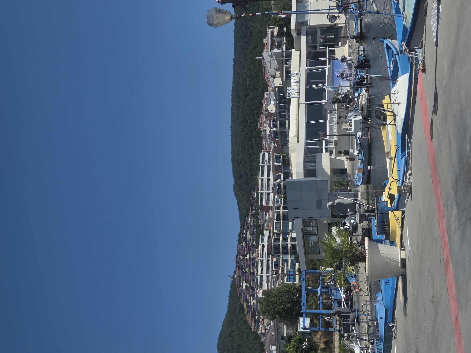 The image depicts a bright, sunny day in the city of Sveti Vlas, Bulgaria, with a clear blue sky dominating the upper portion of the frame. In the foreground, a paved walkway is visible, marked with red and white lines. A variety of small boats, predominantly blue and yellow, are moored in the water. These boats appear to be a mix of dinghies and small motorboats.
Further back, a modern-looking building complex lines the waterfront. This complex features a large, glass-fronted establishment with the words "CODE PRIME" visible on its facade. Several other buildings with balconies and multiple stories rise behind this establishment, with a densely vegetated hillside forming the backdrop. The hillside is dotted with numerous houses or apartments with red-tiled roofs, cascading down the slope. A tall street lamp with an unusual glass fixture stands to the right of the frame, with a flag pole visible nearby. The scene suggests a bustling marina or waterfront area, likely a tourist destination.