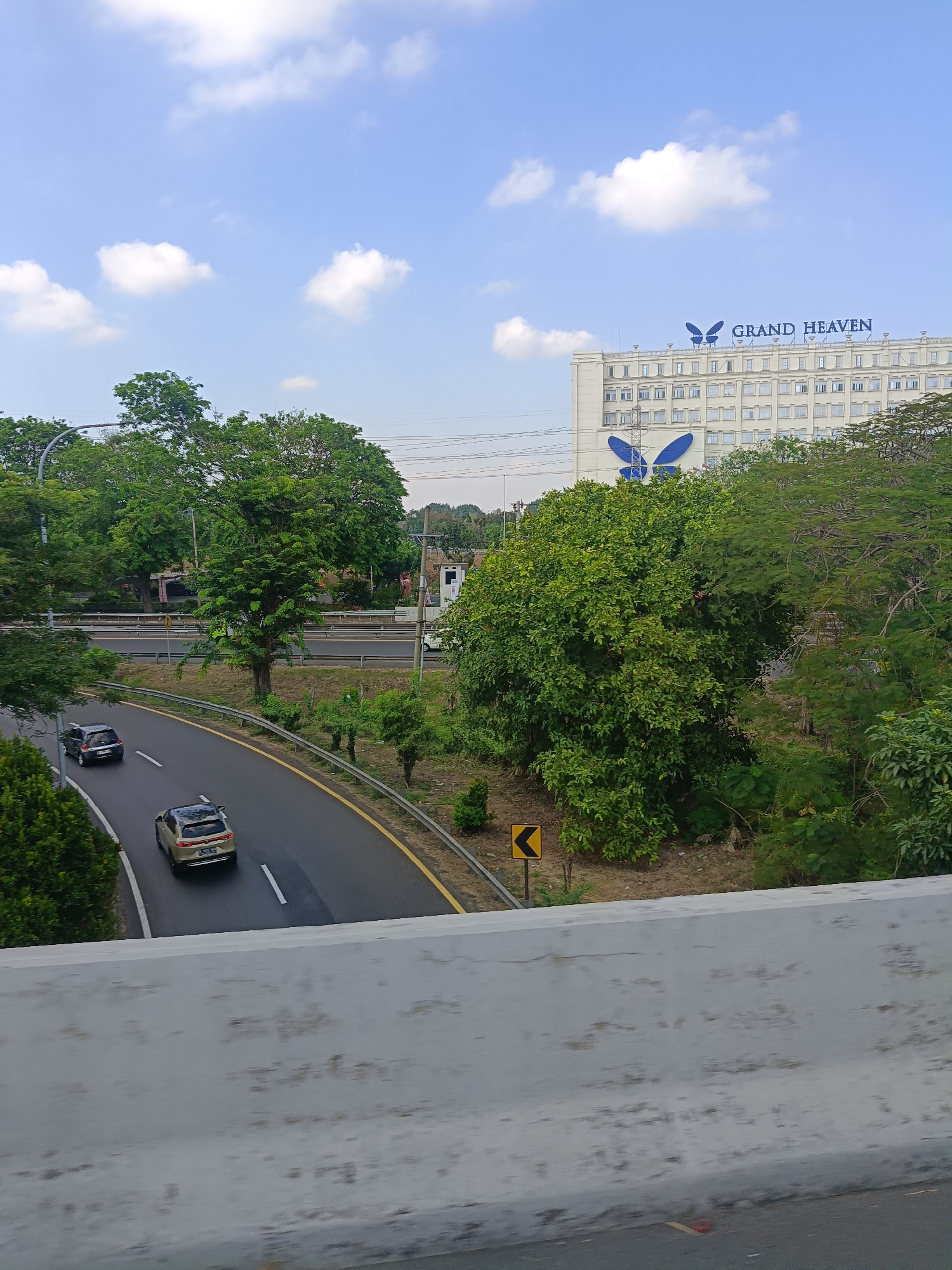 The image captures a daytime view of a highway in Ketegan, Indonesia. A multi-story white building with the words "GRAND HEAVEN" and a large blue butterfly logo is visible in the background. The highway, asphalt with yellow and white lane markings, curves through the scene. Two cars are driving on the highway: a dark-colored car in the left lane and a light-colored SUV in the right lane. Lush green trees and vegetation surround the highway, and a concrete barrier is in the foreground. The sky is blue with scattered white clouds.