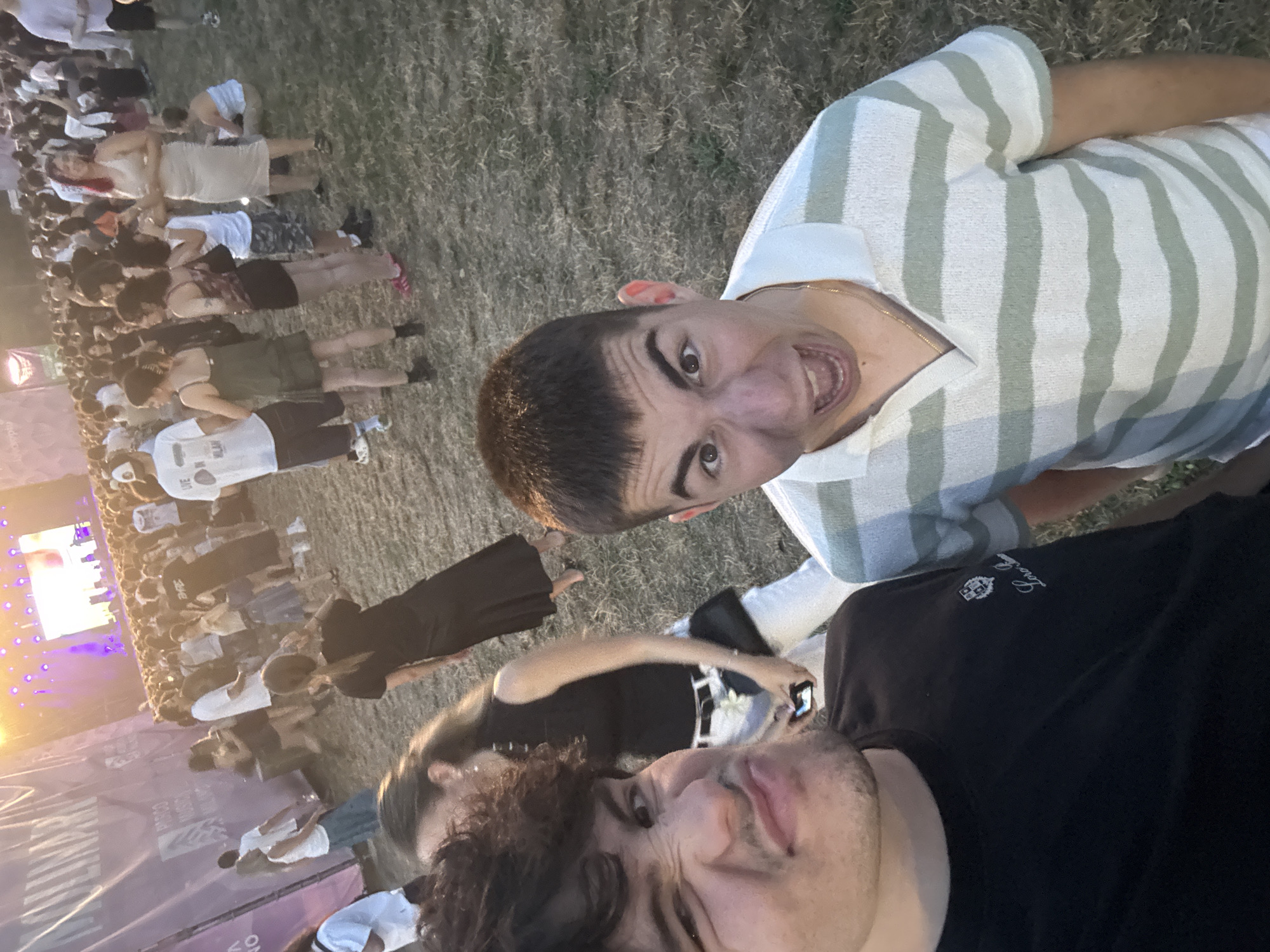 The image captures a crowd of people gathered outdoors, likely at a music festival, given the large stage and lighting visible in the background. The scene is set on a grassy area. Two young men are in the foreground, close to the camera. The man on the left is making a distorted facial expression, while the man on the right is shouting with his mouth open. In the middle ground and background, a diverse crowd is seen milling about, with some individuals dancing and others conversing. Several are holding mobile phones, suggesting they are taking photos or videos. In the far background, a large stage with vibrant purple and white lighting is prominent, with what appears to be performers on it, though they are not clearly discernible. A large banner or screen is also visible to the left, with text partially obscured. The overall atmosphere appears to be energetic and celebratory. The location is identified as Milano Music Week, Parco Musicisti in Segrate, Italy.
