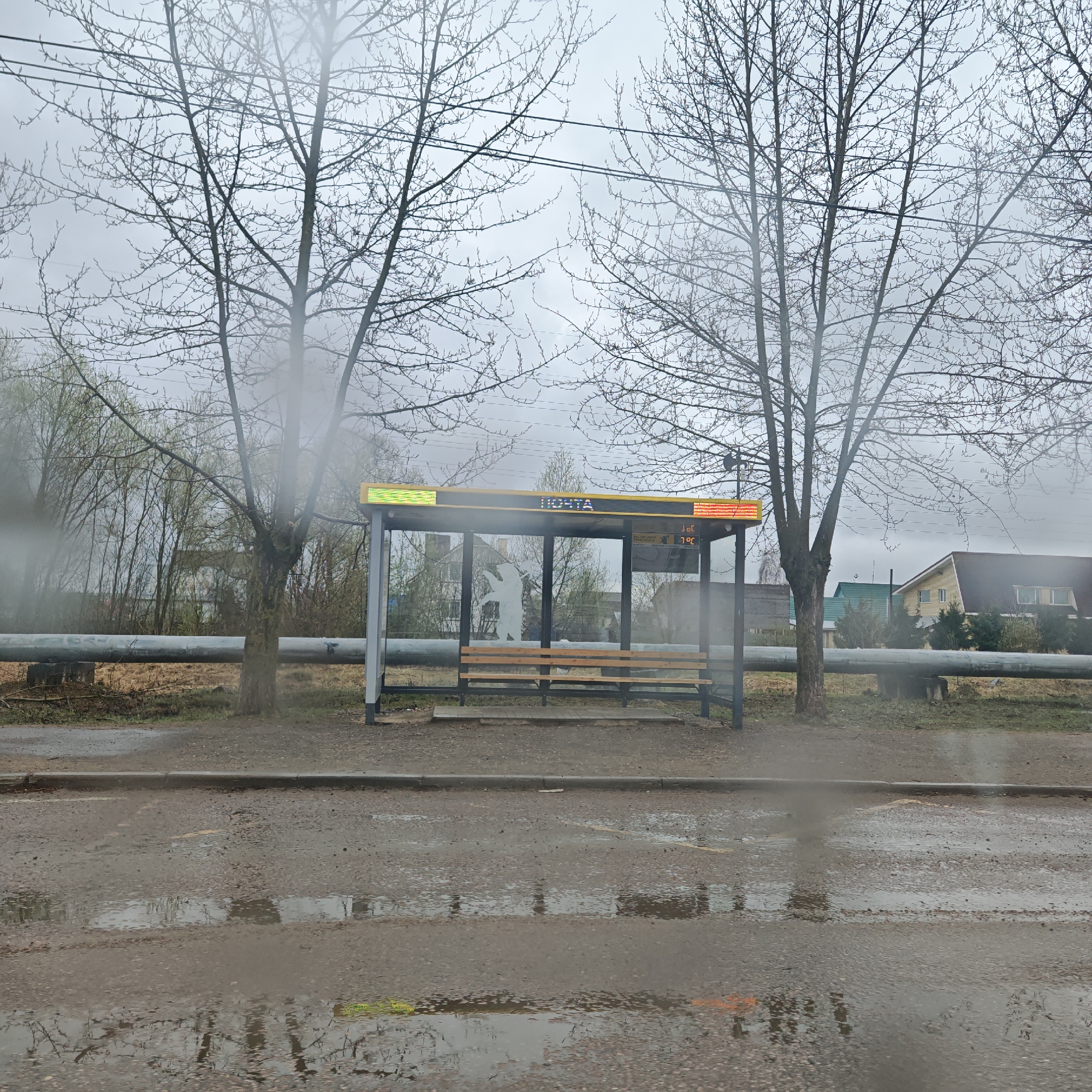 The image depicts a bus stop in what appears to be a rural area of Rostov, Russia. The bus stop is modern, with a digital display on top that reads "ПОЧТА" (Post). Inside the shelter is a simple wooden bench. A white silhouette of a bear is visible on the glass panel of the bus stop. In the background, there are bare trees and buildings, including what appears to be a private house with a brown roof. A large, grey pipe runs horizontally behind the bus stop. The road in the foreground is wet and reflects the scene in its puddles. The overall atmosphere is grey and overcast. The road surface is worn and shows signs of disrepair.