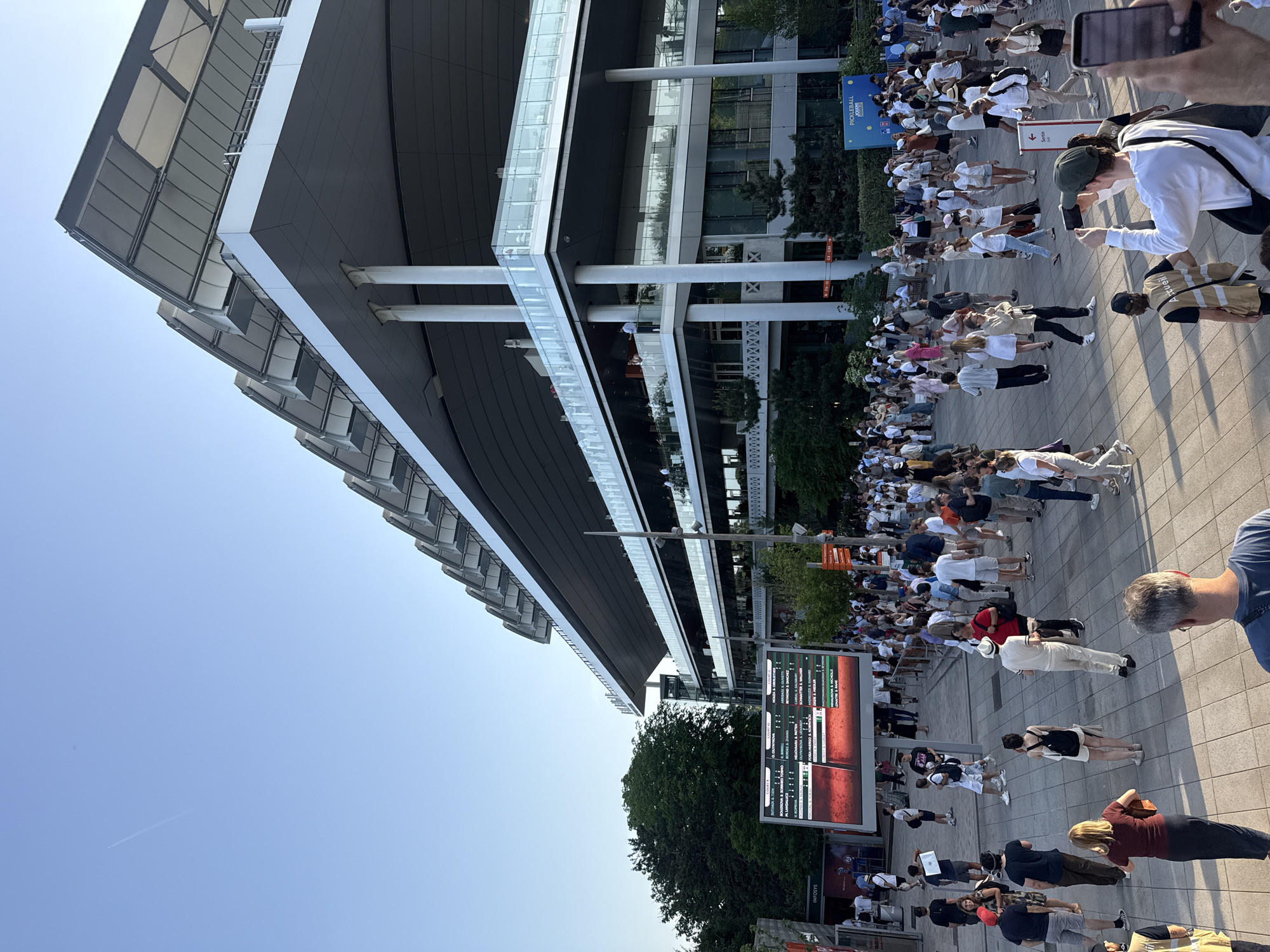The image shows a crowd of people gathered outside a modern building under a clear blue sky. The building has a sleek, angular design with visible architectural elements. A large digital scoreboard displays information, possibly related to a sporting event. The crowd appears to be diverse, with people of various ages and ethnicities. Some individuals are holding up their phones, possibly taking pictures or videos. The setting seems to be a public space, likely an entrance or plaza associated with the building.
Based on the image's elements, this is Roland Garros in Paris, France. The scoreboard shows the results of tennis matches, and the crowd likely consists of spectators attending the tournament.