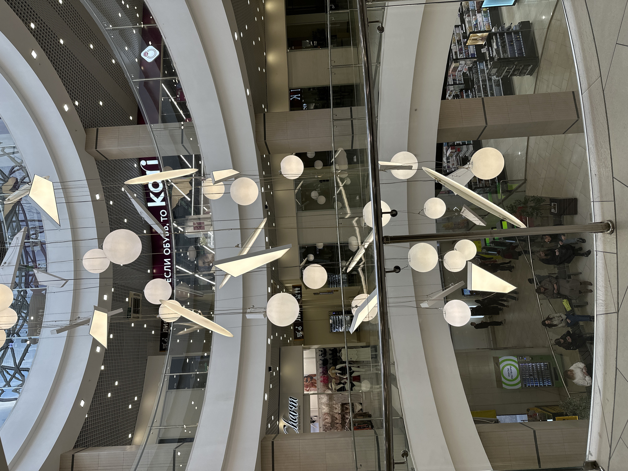 The image captures a high-angle perspective inside a multi-story modern shopping mall in Yekaterinburg, Russia, looking down across various levels. The interior features sweeping, curved white walls, glass balustrades, and a grid-patterned ceiling illuminated by numerous square recessed lights.
Suspended at different heights throughout the central atrium are striking decorative light fixtures, consisting of glowing white spheres and elongated geometric shapes, creating a dynamic visual element. Vertical glass shafts, likely for elevators or design features, reflect sections of the mall.
On the lower visible levels, people are present, some standing and walking, while others are seated on benches, suggesting shopping or resting. These individuals appear as shoppers or mall visitors.
Several commercial signs are visible. A large sign reads "ЕСЛИ ОБУВЬ ТО КА," with another partial view suggesting the full name "Kari," indicating a shoe store. Another store, possibly a lingerie or fashion boutique, displays the name "Charm." On a different level, shelves stocked with cosmetics and perfumes show brand names such as "BURBERRY" and "GIVENCHY." A green vending machine on a lower floor has Russian text that translates to "CHECK YOUR GIFT!" and "MORE PRIZES." Restroom signs ("WC") and a shoe icon are also visible. The lighting is bright and artificial, typical of an indoor commercial environment during daytime.