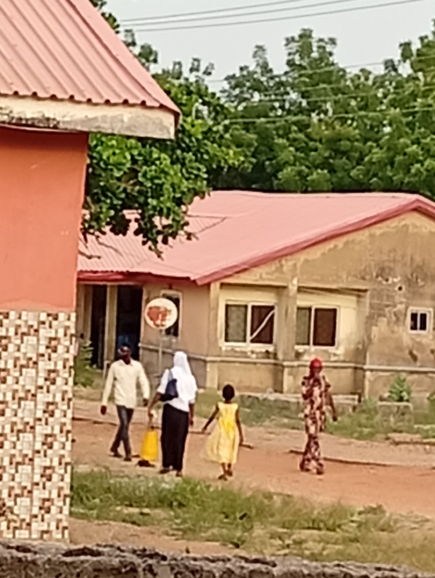 The image depicts a scene in what appears to be a residential or community area. Several people are walking on a dirt path between buildings with reddish roofs. A man in a light-colored shirt walks with a woman in a white headscarf and dark skirt, who is holding a yellow object. A girl in a yellow dress walks next to them. Another woman in a patterned dress walks further away on the path. The building behind them has a satellite dish affixed to the exterior. The sky is overcast and trees are visible in the background. A building with reddish-brown and tile-patterned walls is partially visible on the left. The location is in the city of Unknown, Unknown.