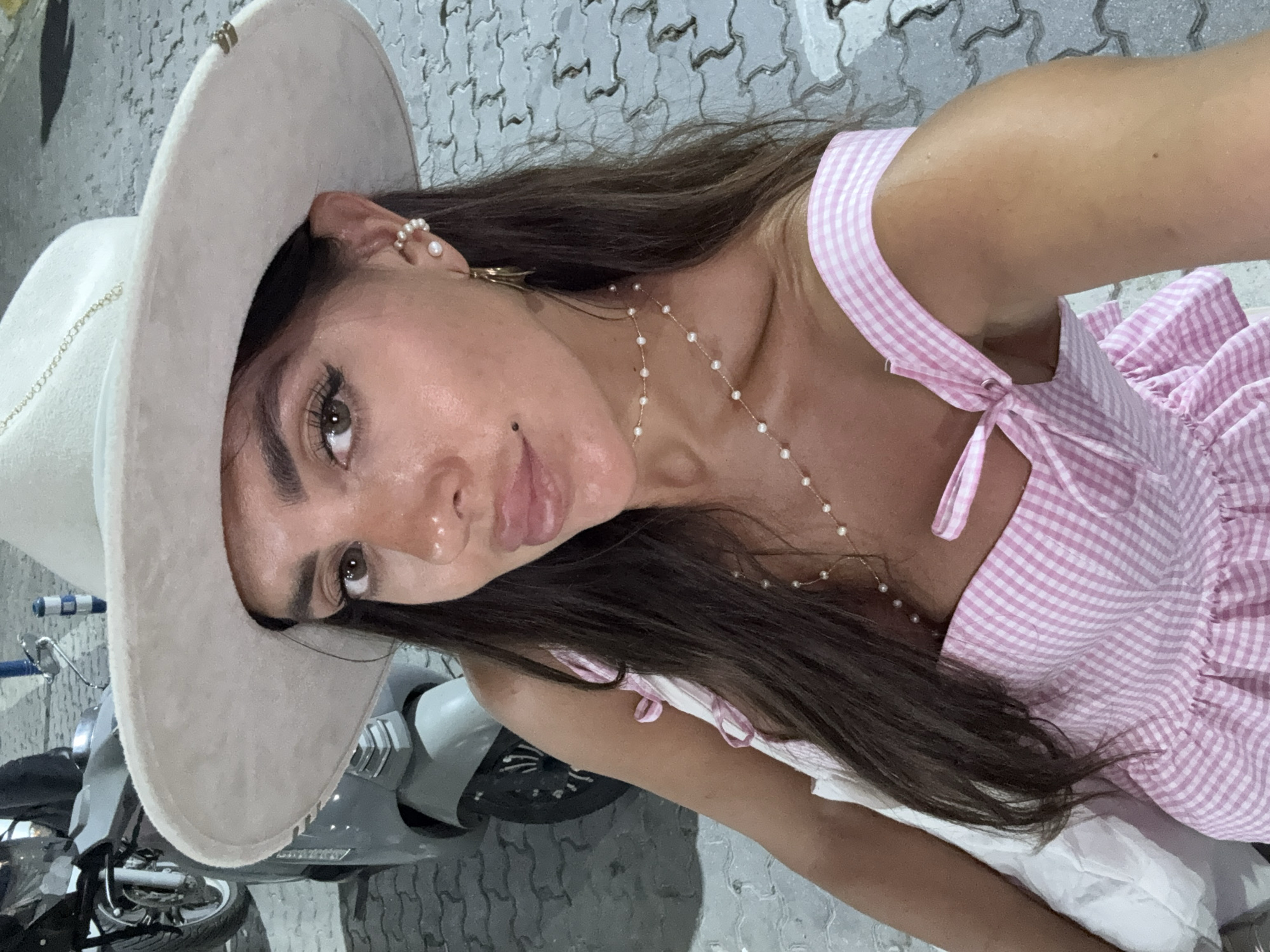 A woman wearing a wide-brimmed hat and a pink and white gingham dress is captured in a selfie. She has long dark hair, brown eyes, and is wearing pearl earrings and a pearl necklace. Her lips are slightly parted. The background shows a cobblestone ground, part of a scooter, and what appears to be a street in Fethiye, Turkey, possibly near a harbor or waterfront area, suggested by the scooter and paving stones.