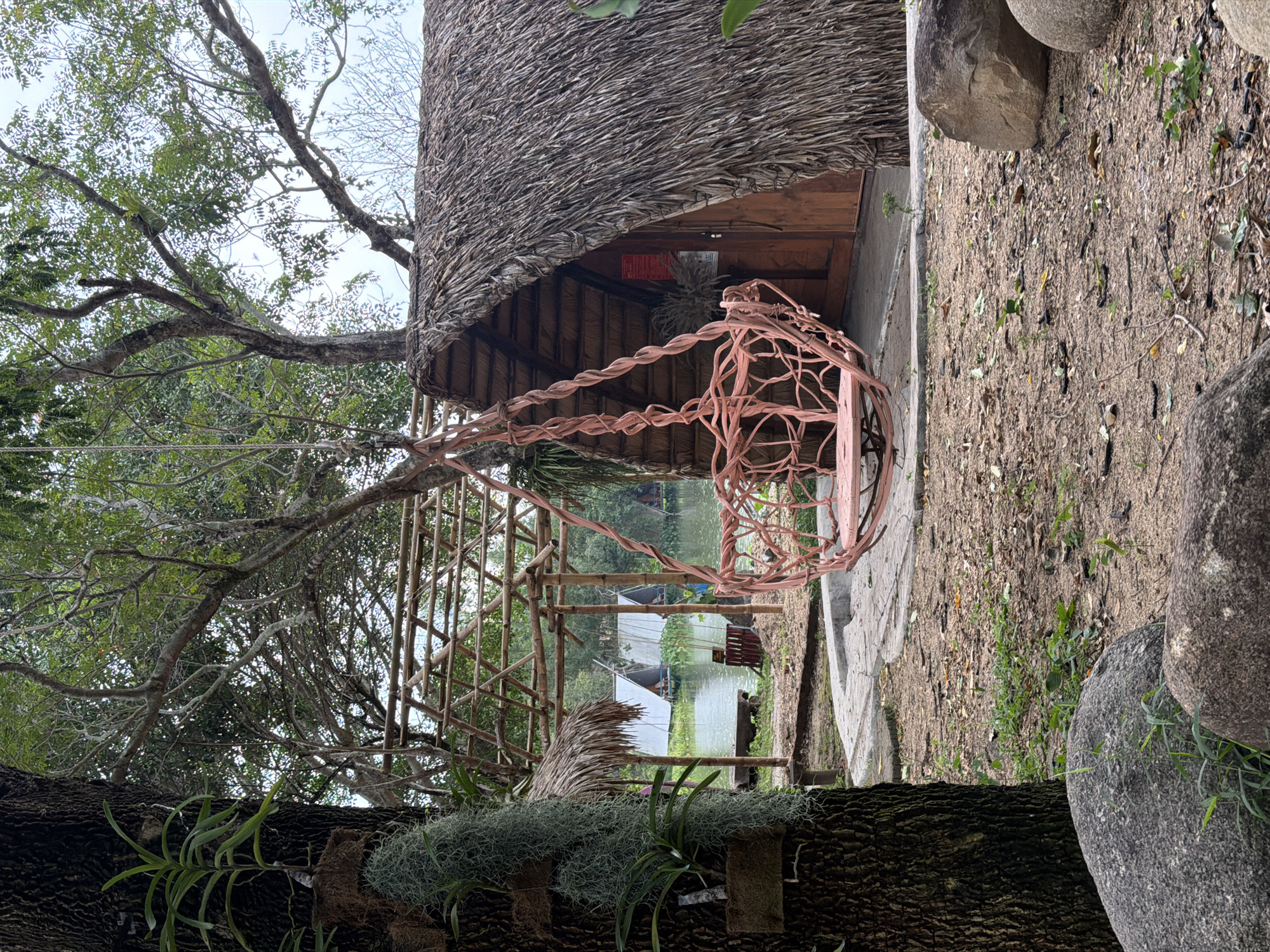 The image depicts an outdoor scene in Khánh Vĩnh, Vietnam, characterized by a rustic, natural setting. In the foreground, a large tree trunk with textured bark is prominent on the left, adorned with hanging plants and epiphytes. A pink, woven swing chair hangs from a tree branch, positioned slightly to the right of center. Behind the swing, a small, A-frame hut with a thatched roof and wooden door is visible. To the left of the hut, a bamboo structure with a thatched roof extends over a path leading towards a body of water. In the background, the water is dotted with several white tent-like structures on rafts or boats, suggesting a resort or accommodation area. The ground is covered with dirt, fallen leaves, and some scattered stones, indicating a natural, unpaved area. Lush green foliage and trees frame the scene, contributing to the serene, natural ambiance.