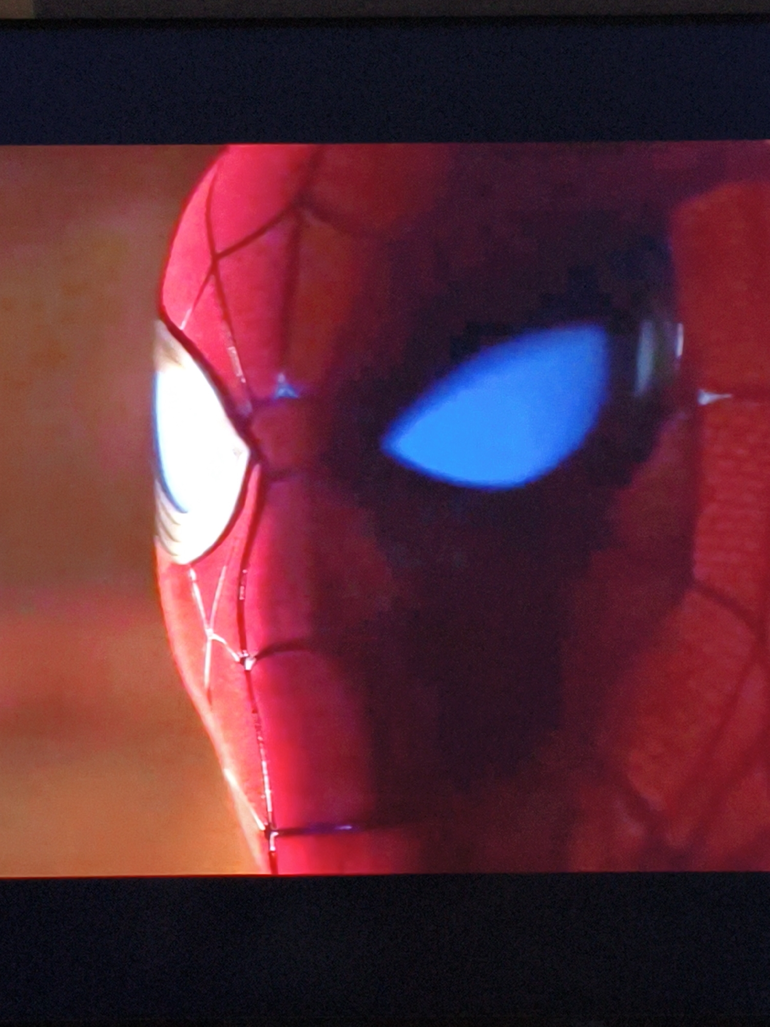 The image shows a close-up of the Spider-Man mask. The mask is predominantly red with white webbing details. The left eye lens is bright white, reflecting light, while the right eye lens is a vivid blue. The background is blurred, with warm, orangey-red tones, suggesting an indoor or dimly lit environment, possibly a movie screen or a photograph. There is no indication of the city of Vladivostok, Russia, or any specific location within it.