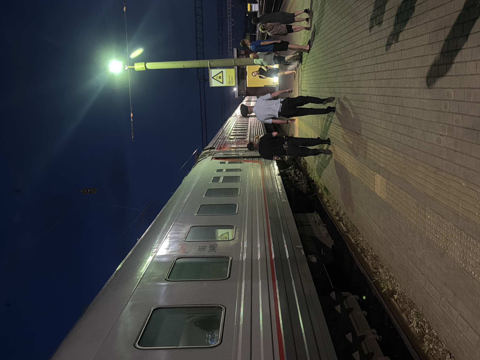 At night, a train with a silver-grey exterior and red accents is parked at a station platform. The train has multiple windows, some reflecting light. On the platform, two individuals in dark uniforms and hats are walking away from the camera, towards the train. Further down the platform, several other people are standing and walking, including a woman in a dark dress, a man in a light blue t-shirt, a child in a pink and grey patterned shirt, and two other individuals. A yellow lamppost with a bright light is prominently featured, casting light on the scene. A yellow triangular sign with a pictogram of a train is affixed to the lamppost. The platform is paved with light-colored bricks, and there is grass and debris along the edge of the train tracks. The sky is dark blue, indicating nighttime.