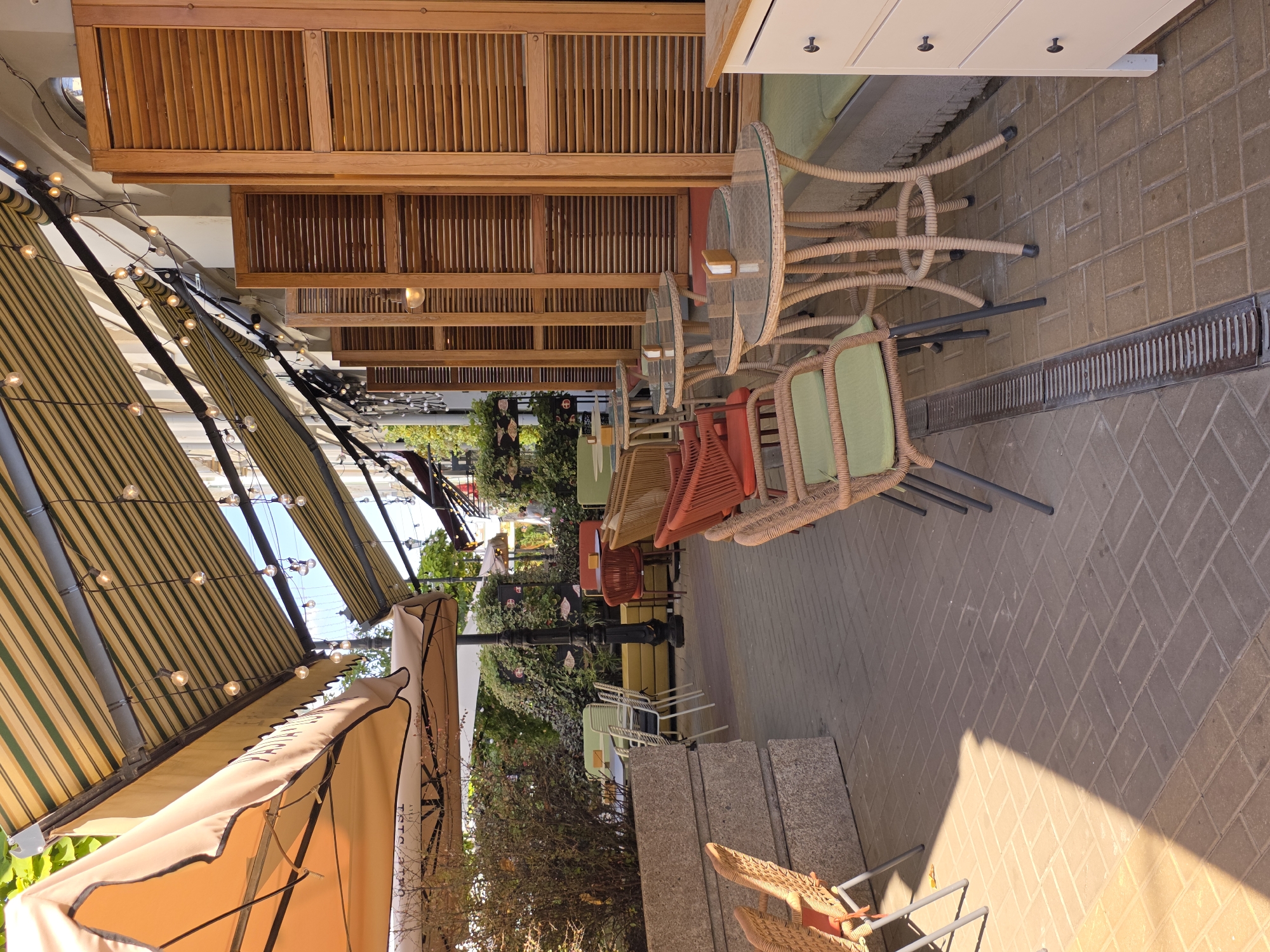 The image depicts the outdoor seating area of a cafe located in Rostov-on-Don, Russia. The cafe features multiple tables and chairs arranged on a brick-paved patio. Overhead, striped awnings provide shade, and string lights are hung between them. The tables are round with glass tops and rattan bases. The chairs are also made of rattan with light green cushions. To the right, a white cabinet with dark handles is visible. In the background, there is lush greenery and some buildings. The scene is empty of people, suggesting the cafe is either closed or not yet busy. The overall impression is that of a pleasant, sunny day at a cafe.