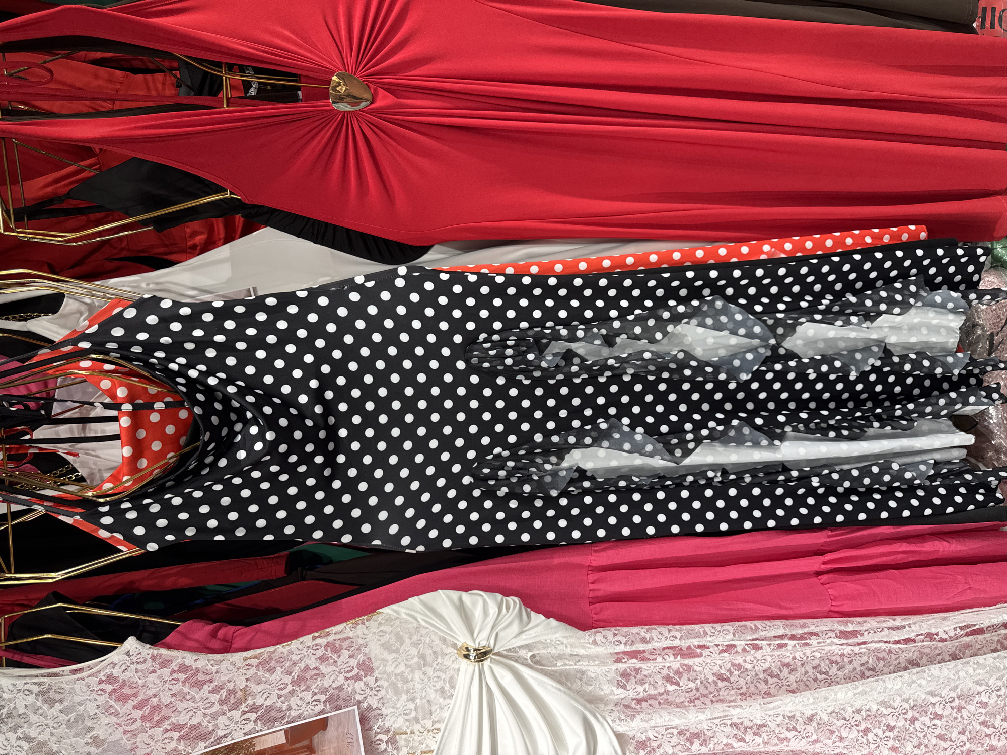 The image depicts a close-up view of several dresses hanging on a rack, likely in a store in Gungoren, Turkey. The foreground is dominated by a black dress with white polka dots, featuring ruffled accents and a halter neckline. To the left of this dress, a white lace dress is visible, partially obscured by a bright pink dress. To the right of the polka dot dress, a vibrant red halter dress with a gold heart-shaped embellishment at the bust is prominent. The dresses are arranged closely together, suggesting a retail display. The background shows glimpses of other clothing items and the metallic structure of the clothing rack. The lighting appears to be artificial, common in indoor retail settings.