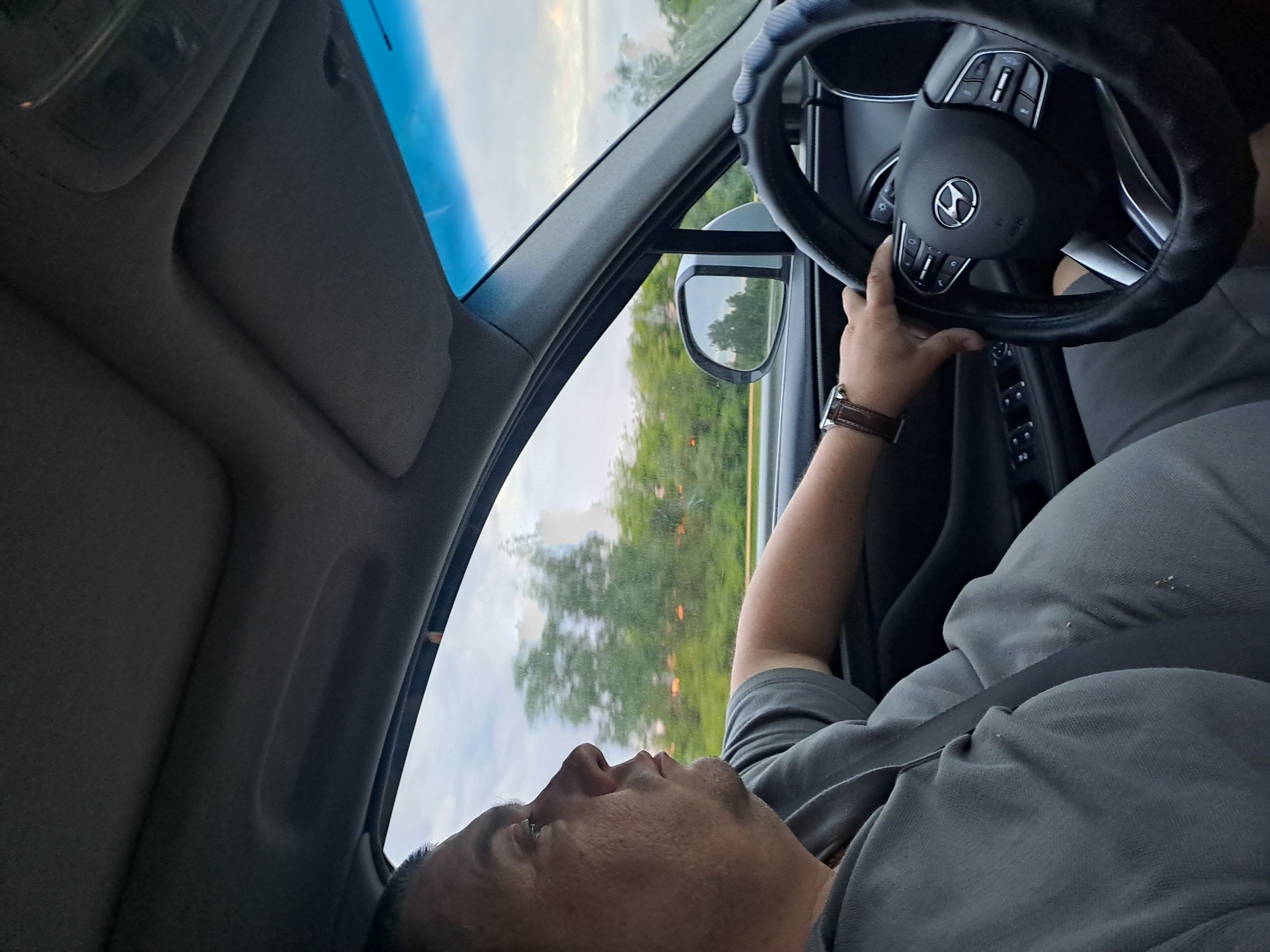The image depicts the interior of a car, focusing on the driver from a passenger's perspective. The original image is rotated 90 degrees counter-clockwise. A person, presumably male, is seated in the driver's seat, wearing a gray t-shirt and a seatbelt across their chest. A brown leather-strapped watch is visible on their left wrist, which rests on the steering wheel. The driver's face is seen in profile, looking forward and slightly upwards, with a neutral expression.
The steering wheel, featuring a black cover for grip, has a prominent Hyundai logo in its center, indicating the car's make. Various control buttons are visible on the steering wheel spokes. Through the car windows, a natural landscape of green trees and an overcast or partially cloudy sky can be seen, suggesting the vehicle is in motion on a road or highway. The driver's side rearview mirror reflects the passing scenery. The car's interior, including the dashboard and overhead lining, appears standard.
The activity taking place is driving. The time of day is ambiguous due to the cloudy sky, but it is daylight. No specific location cues beyond the natural landscape are visible to pinpoint the exact location within Mayak, Russia. No other significant text apart from the car's emblem is discernible.