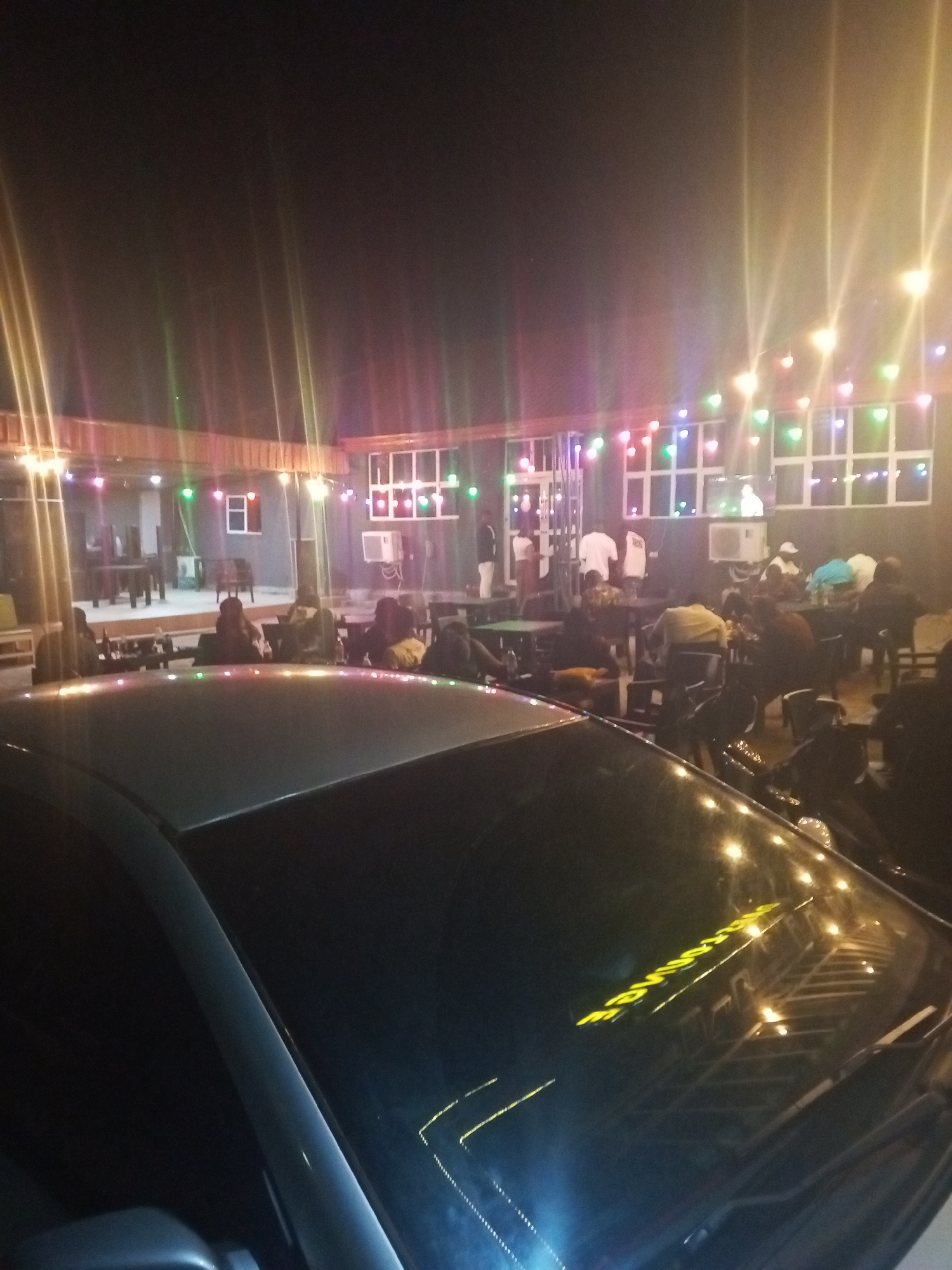 This nighttime photograph captures a bustling outdoor or semi-outdoor restaurant or bar scene in Lokoja, Nigeria. The setting is brightly lit by numerous colorful string lights, which are draped across an overhead structure and visible in reflections, creating a festive and social atmosphere.
Many people are present, seated at tables in groups, suggesting they are dining, socializing, and relaxing. Some individuals are also seen standing within the establishment. The background features a building with prominent white-framed windows, some of which also display colorful lights.
In the foreground, the hood and windshield of a dark-colored car are visible, partially obscuring the view of the patrons. The car's surface reflects the overhead string lights. On the car's windshield or roof, there is an inverted yellow reflection of text. Although partially obscured and reversed, a segment of this text appears to read "SSURINE E," which could be part of a sign from the establishment or a nearby business. The dark sky confirms the late time of day, highlighting the artificial illumination that defines the space. The overall impression is that of a popular and lively evening gathering spot.