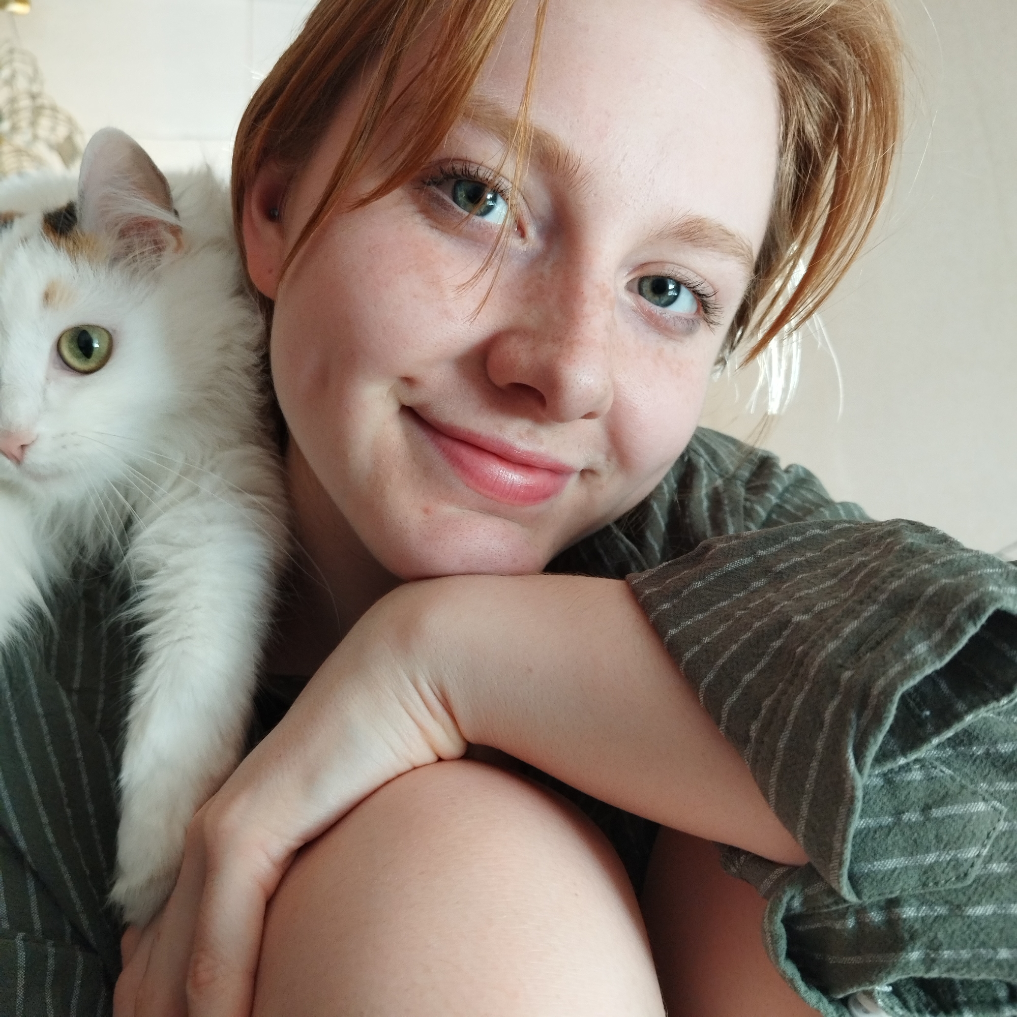 A close-up, selfie-style image shows a young woman with red hair and fair skin smiling at the camera. She has freckles on her face and green eyes. A white cat with some tan markings is resting on her left shoulder, looking to the left. The woman is wearing a striped green shirt. Her arm rests on her knee in the foreground. The background appears to be a neutral, light-colored wall. No specific location in Yekaterinburg, Russia, is identifiable from the image.